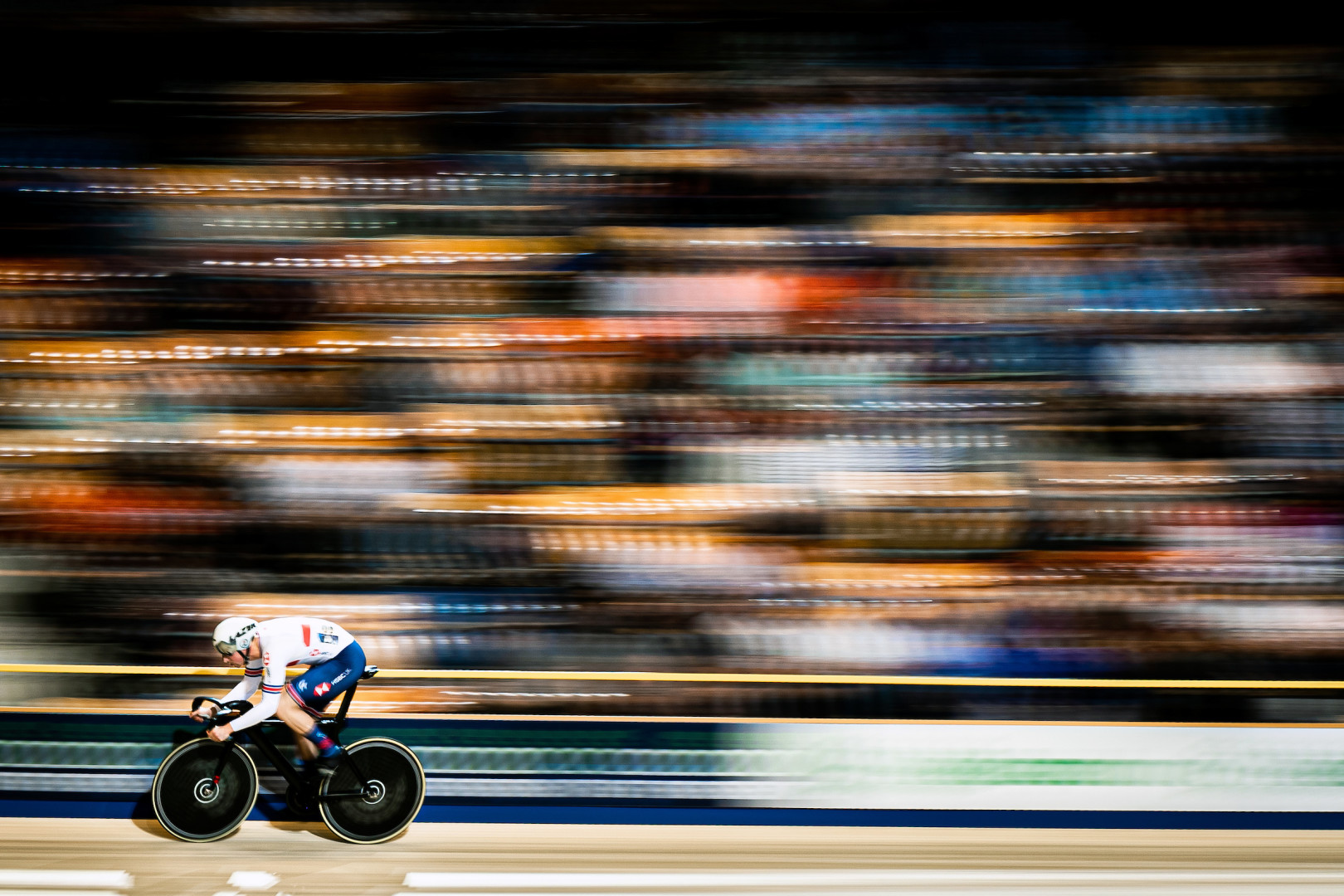 Cyclist flying around the velodrome track as crowd blurs into colours.