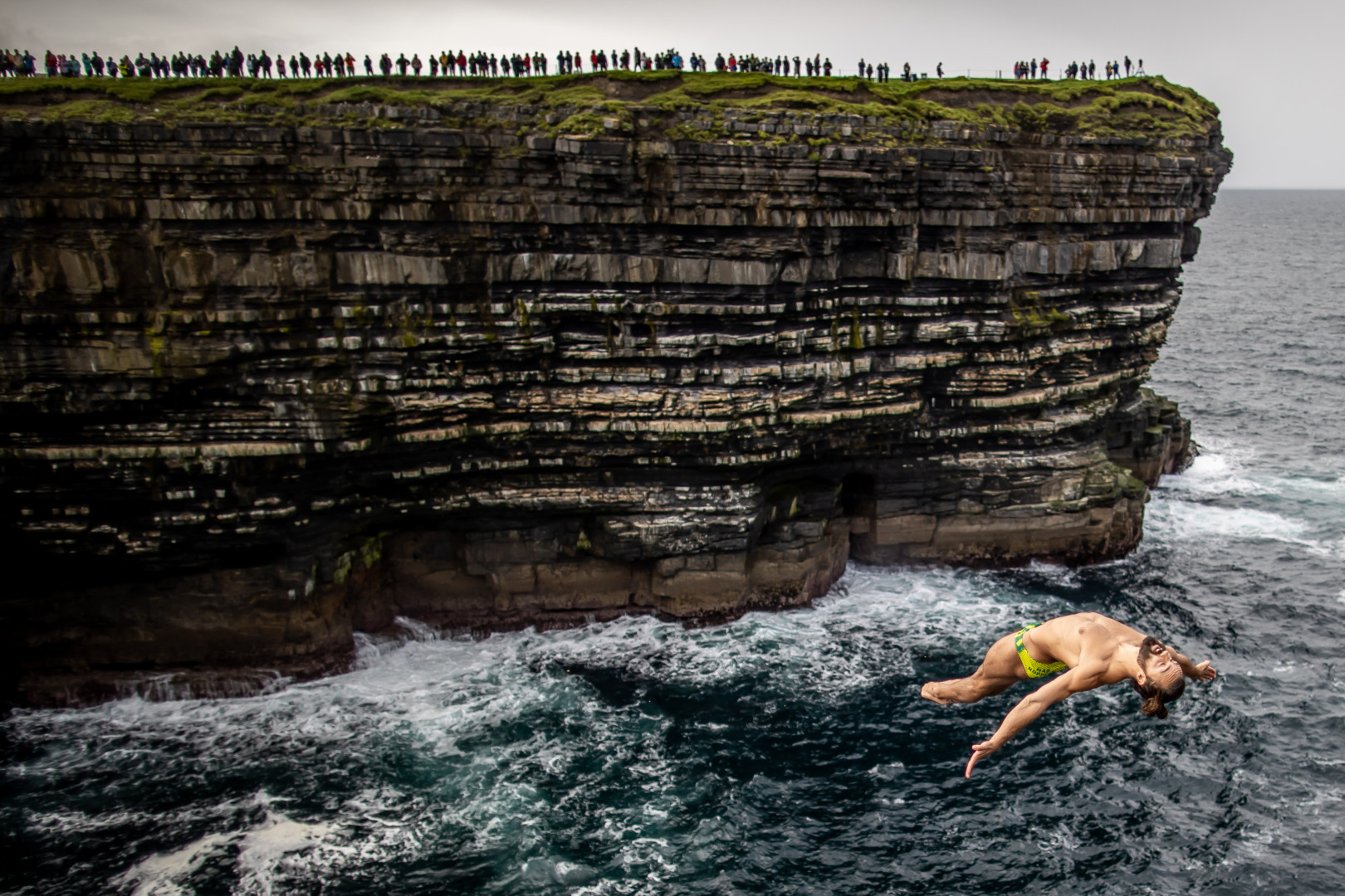 Male cliff diver mid air whilst onlookers watch from distant cliff.