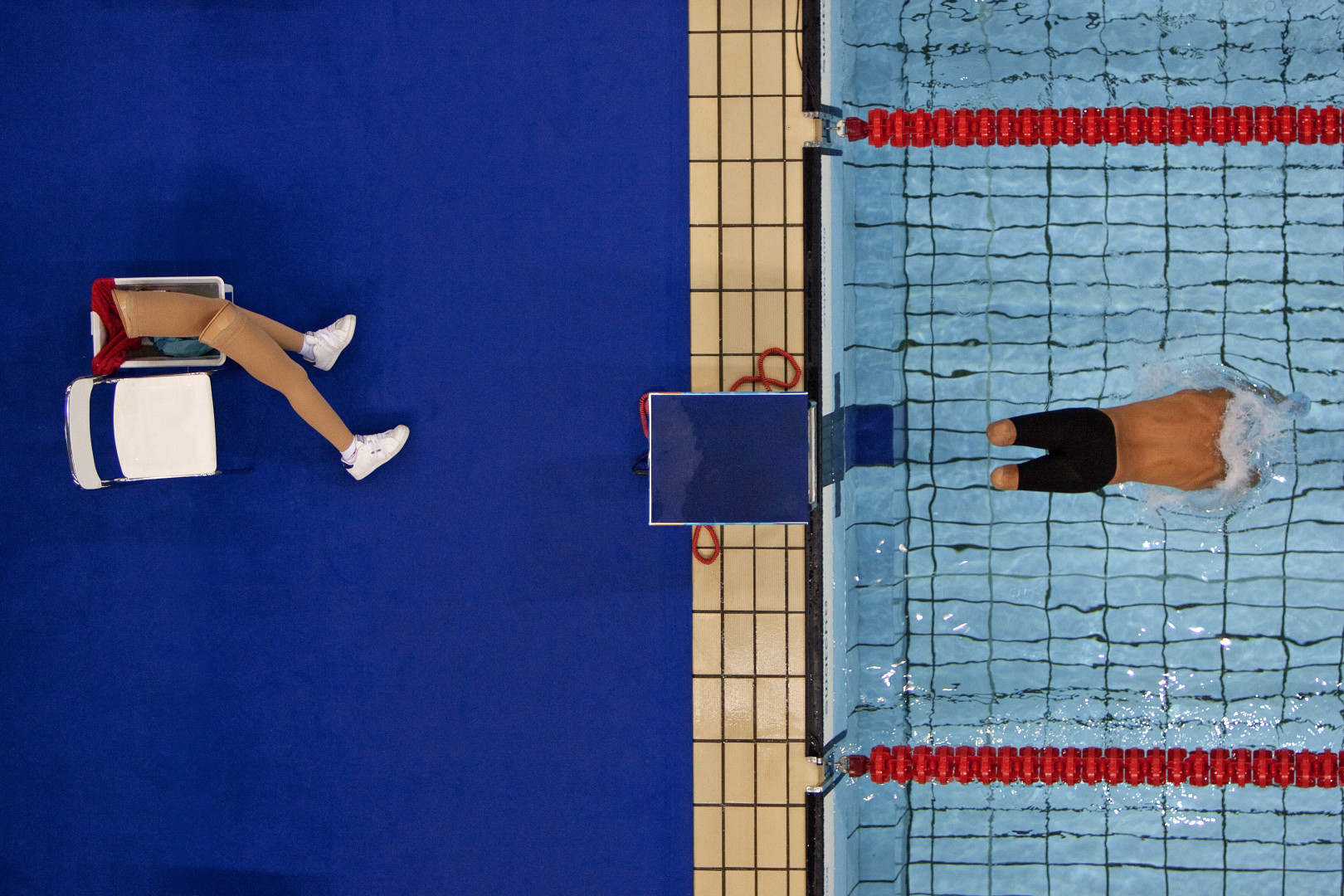 Paralympic swimmer mid diving into pool as prosthetic legs sit on the side of the pool.