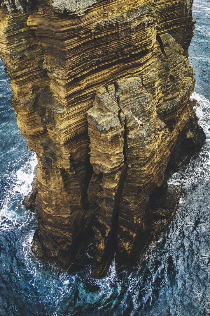 Tiny diver mid air diving off of huge cliff into the sea.