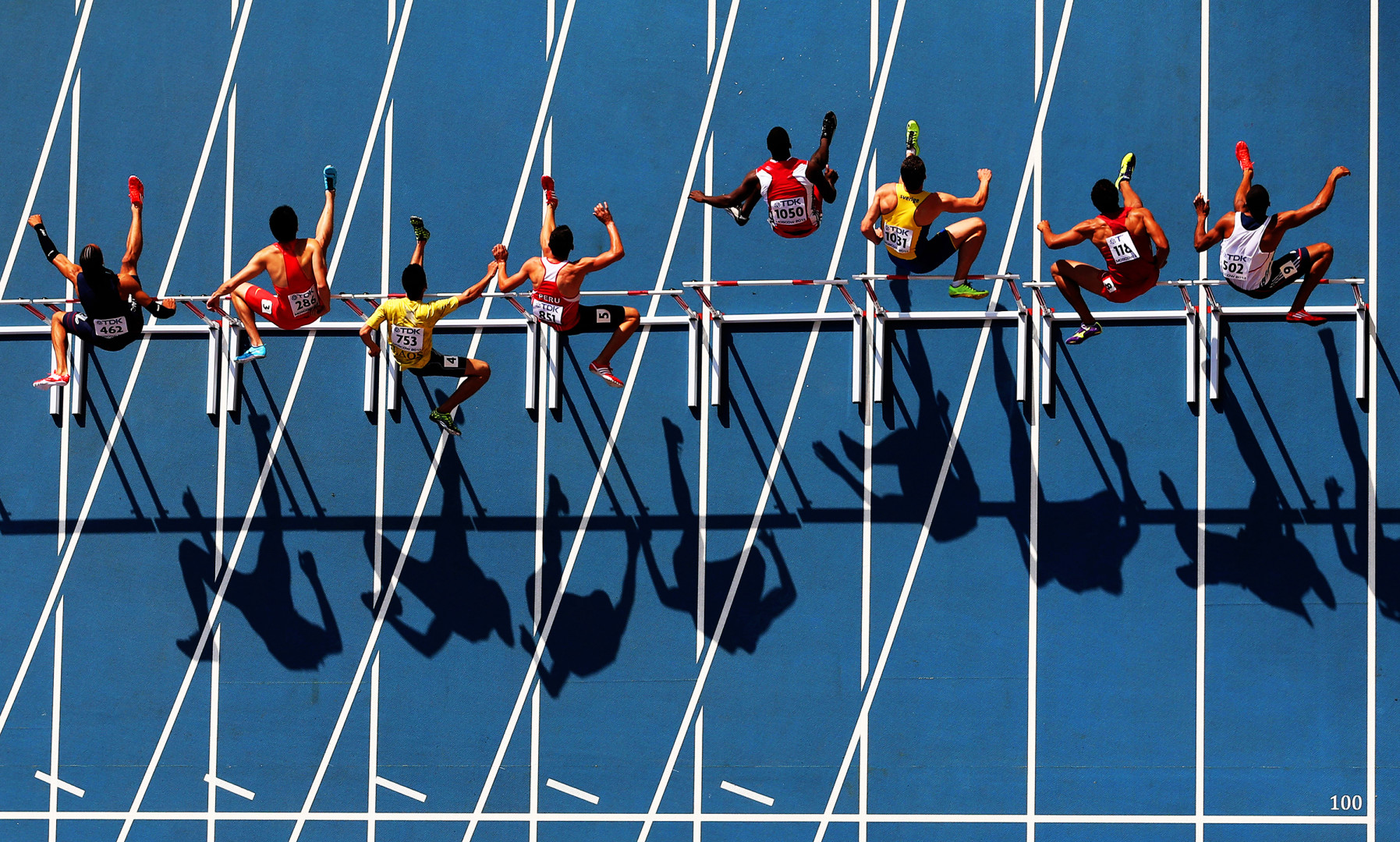 Birdseye view photograph of athletes mid jump over hurdles on blue running track.