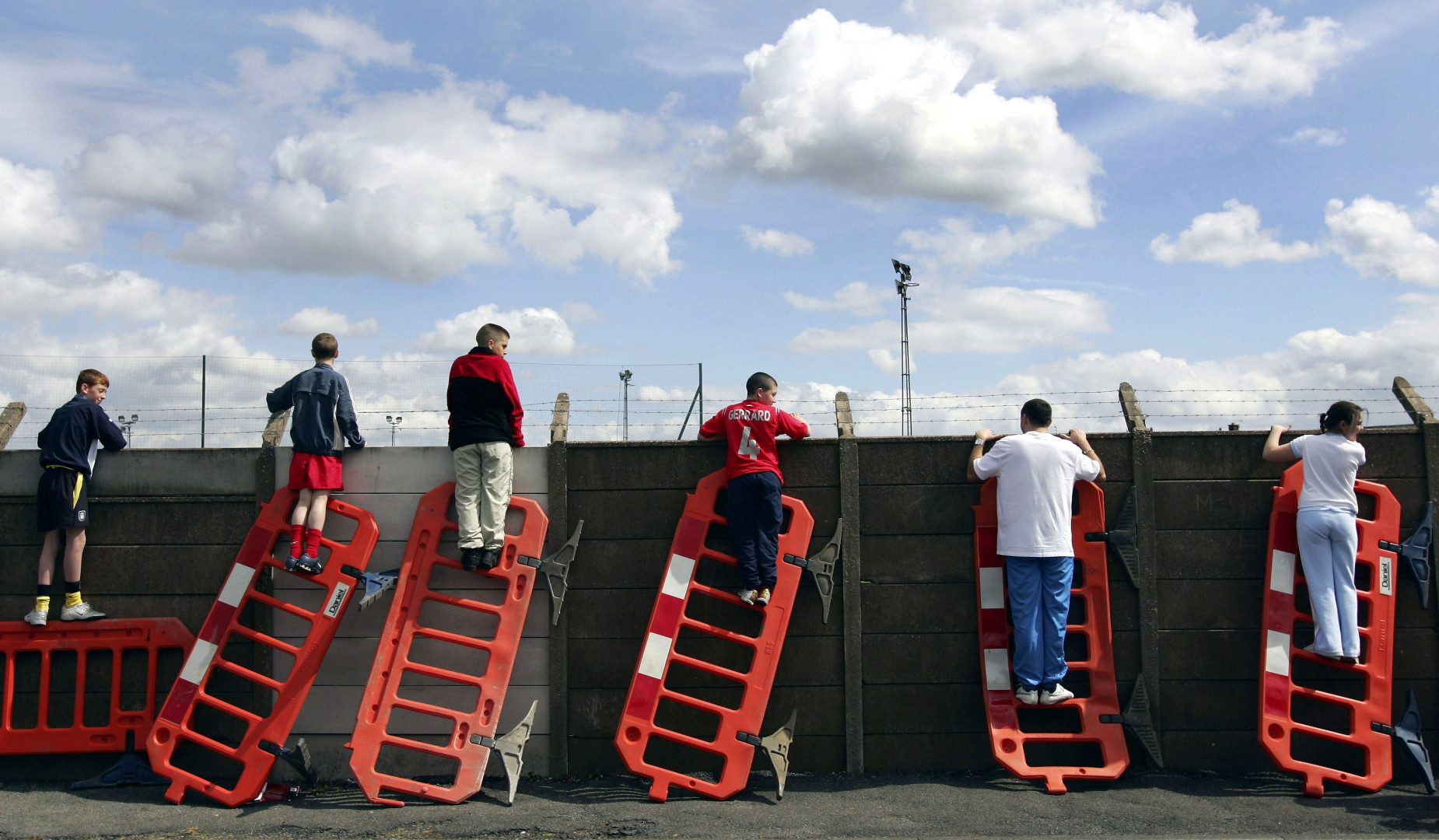 Young football fans using road-work fences as ladders to look over the wall and watch a football match.