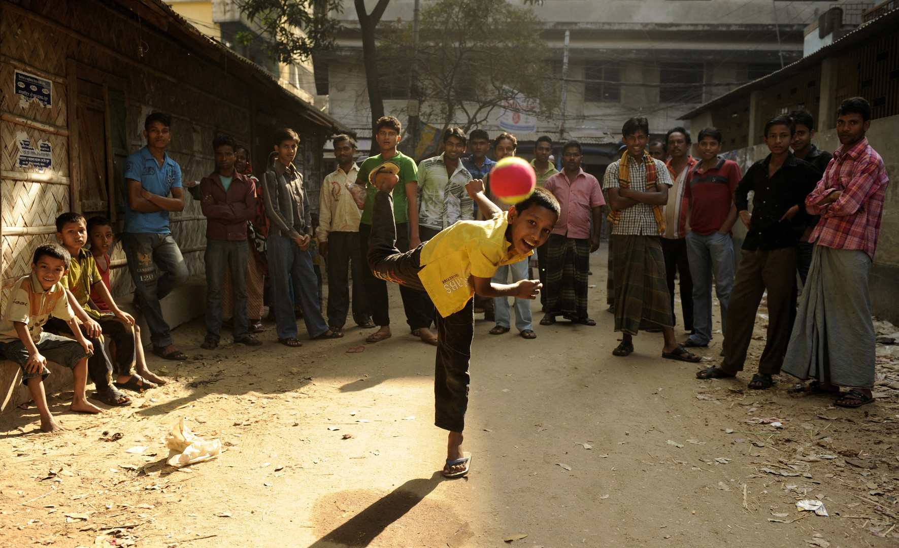 Young Chittagong boy throwing ball at the camera lens as onlookers watch.
