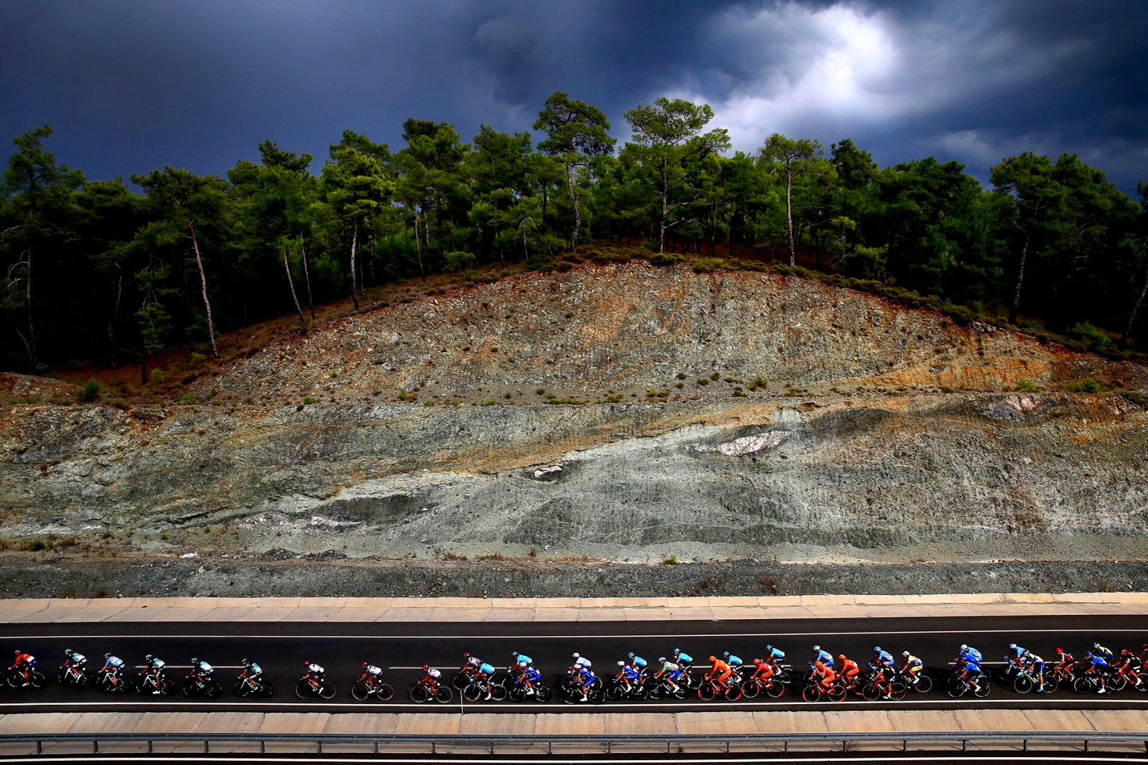 Tiny cyclists cycling along the road below a cliff and under stormy weather.