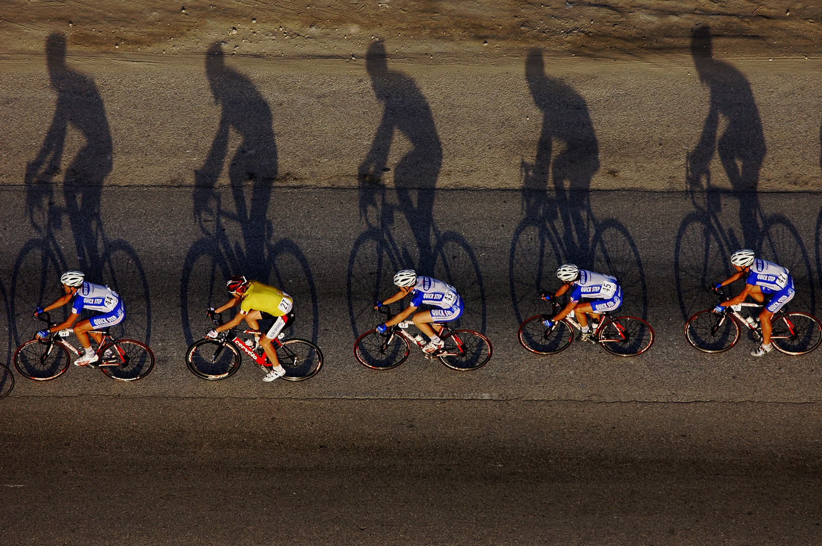 Overhead photograph of a line of cyclists, as their shadows stretch out across the road.
