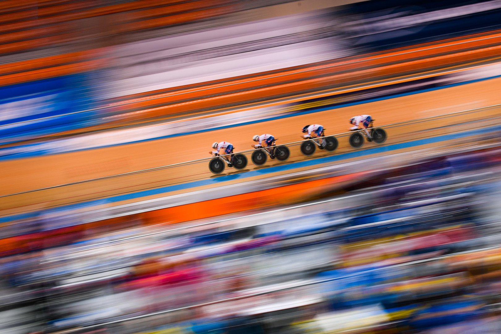 Diagonal photograph of England Cyclists in focus, riding full speed around Velodrome track, as the crowd blur into colour and movement.