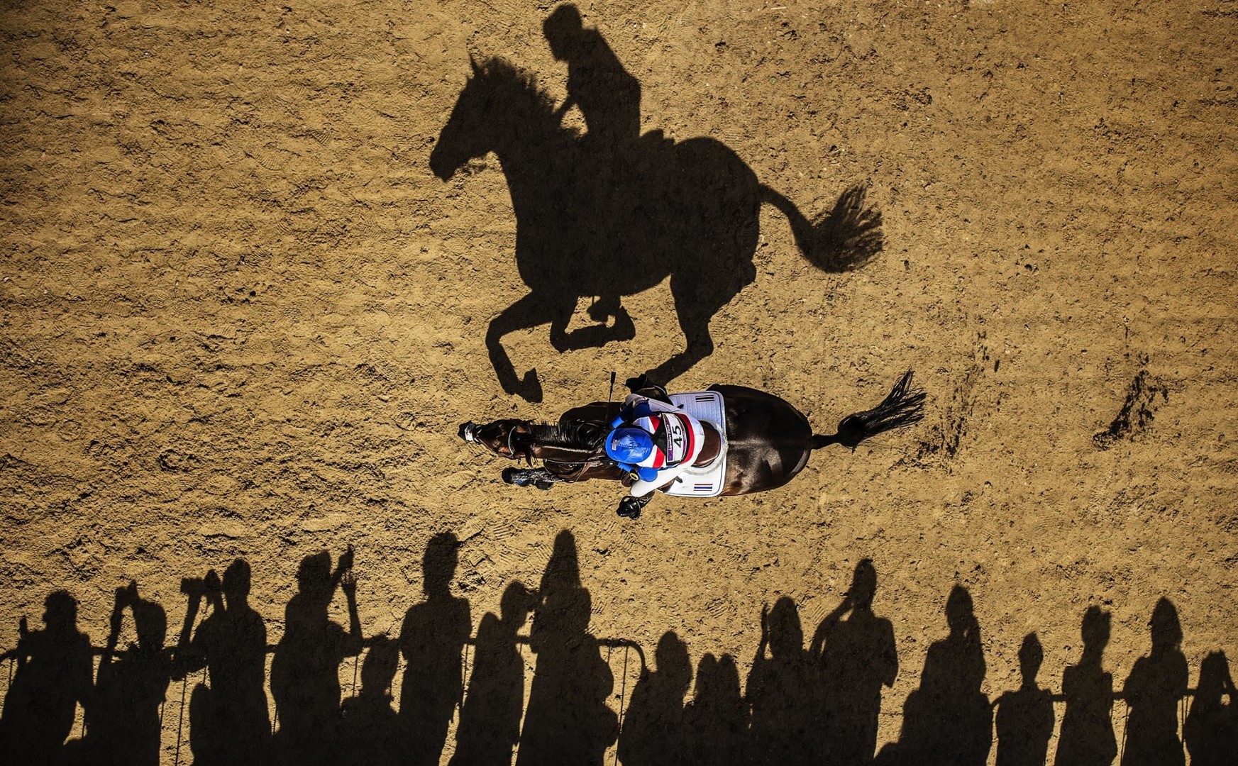 Birdseye shot of jockey and horse during a cross country race with shadows of the crowd, jockey and horse reflecting on the track.