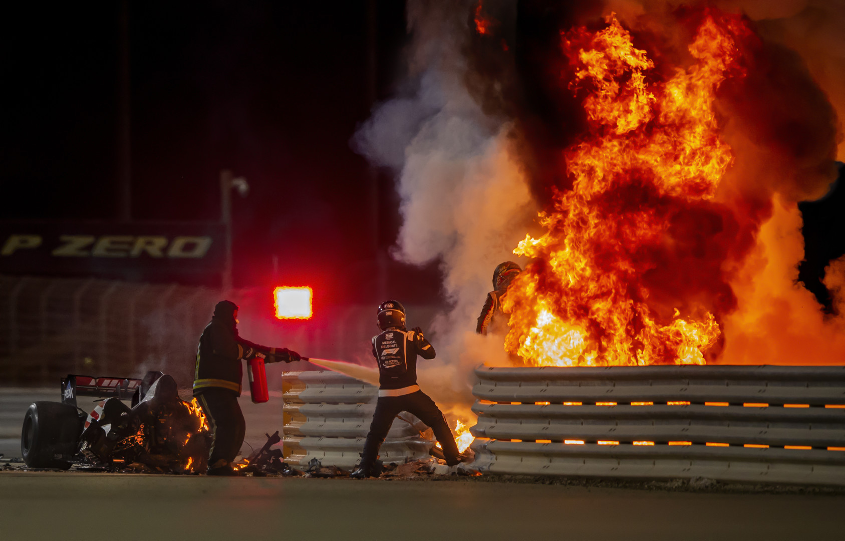 Formula 1 driver and safety officers extinguish burning vehicle as Romain Grosjean emerges from flames.
