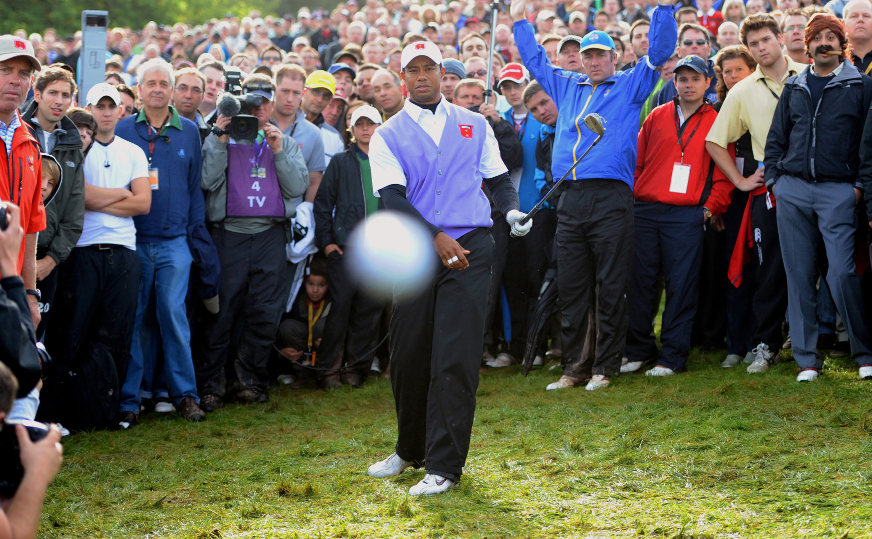 Tiger Woods and crowd watching on as golf ball flys towards camera lens.