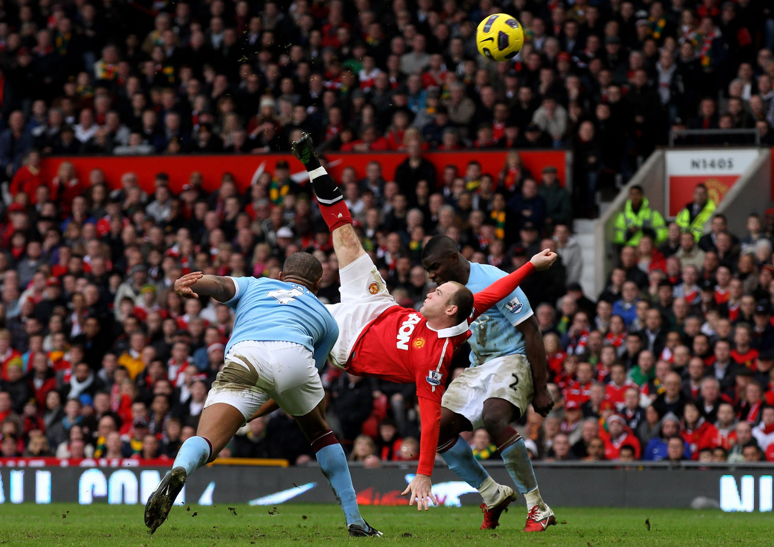Wayne Rooney taking an over-head  kick in between two Manchester City players.