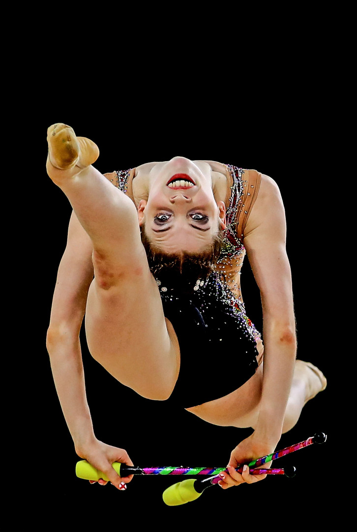 Rhythmic gymnast mid air in splits jump as her head is flung backwards looking directly at the camera.