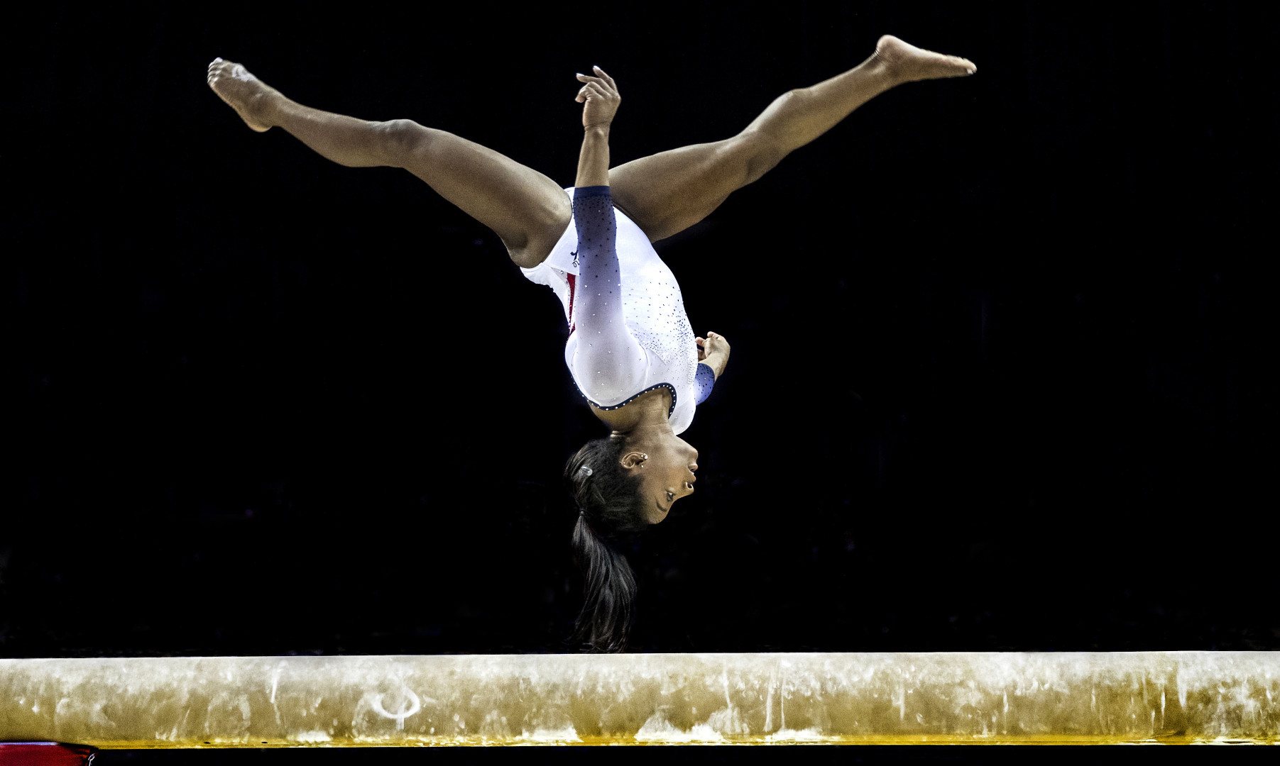 Simone Biles upside down whilst performing a cartwheel jump on a balance beam.