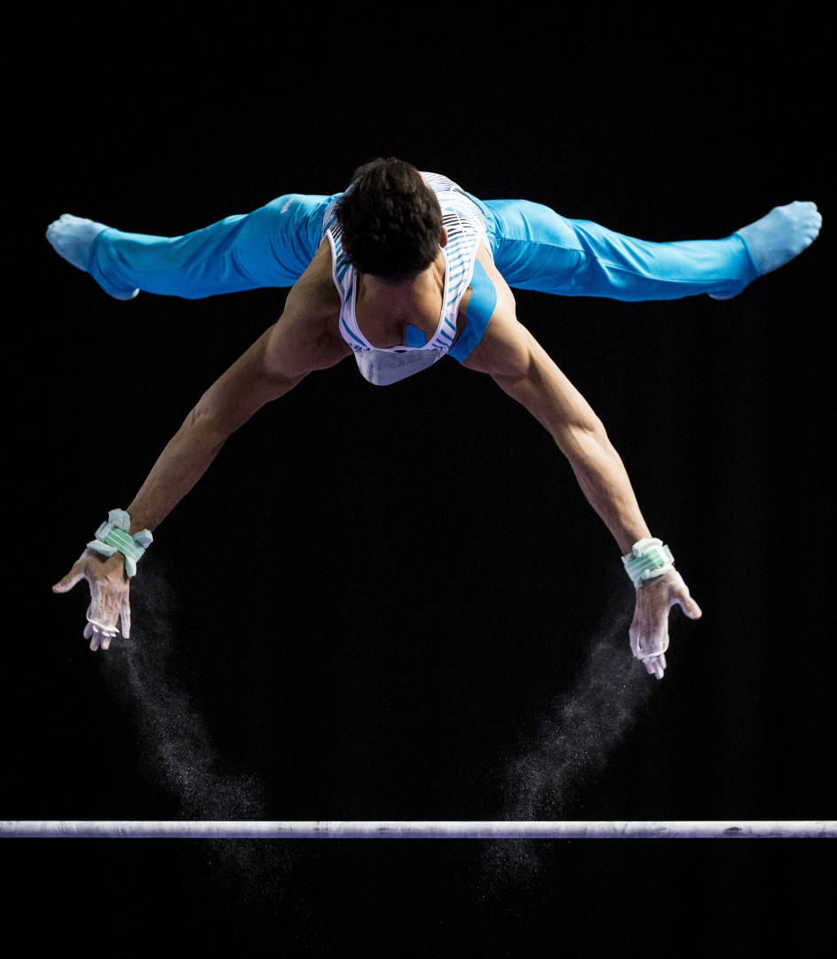 Male gymnast mid air in an 'X' position as he launches himself from the swing bars.