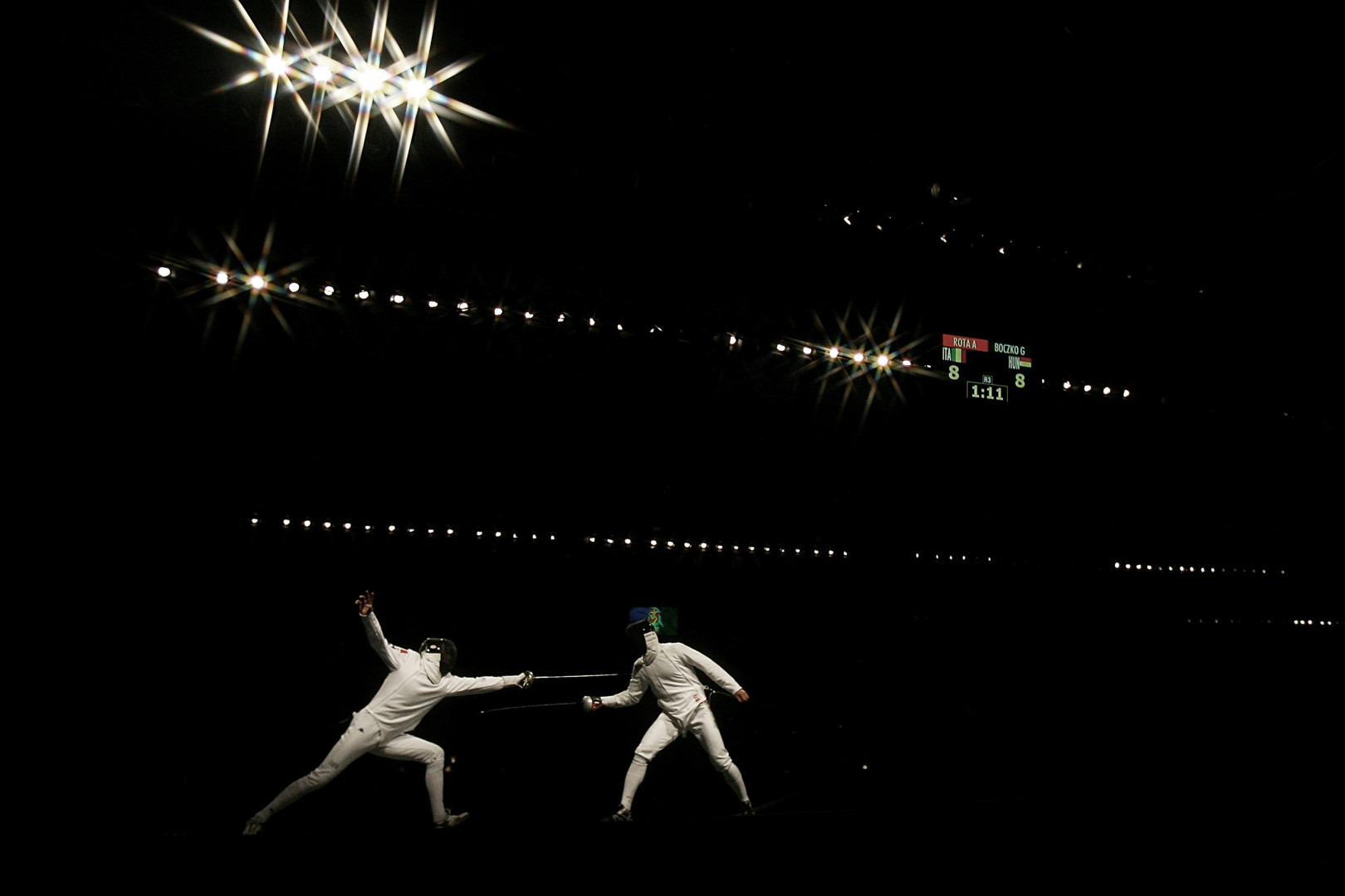 Two athletes fencing whilst spotlighted in their white outfits.
