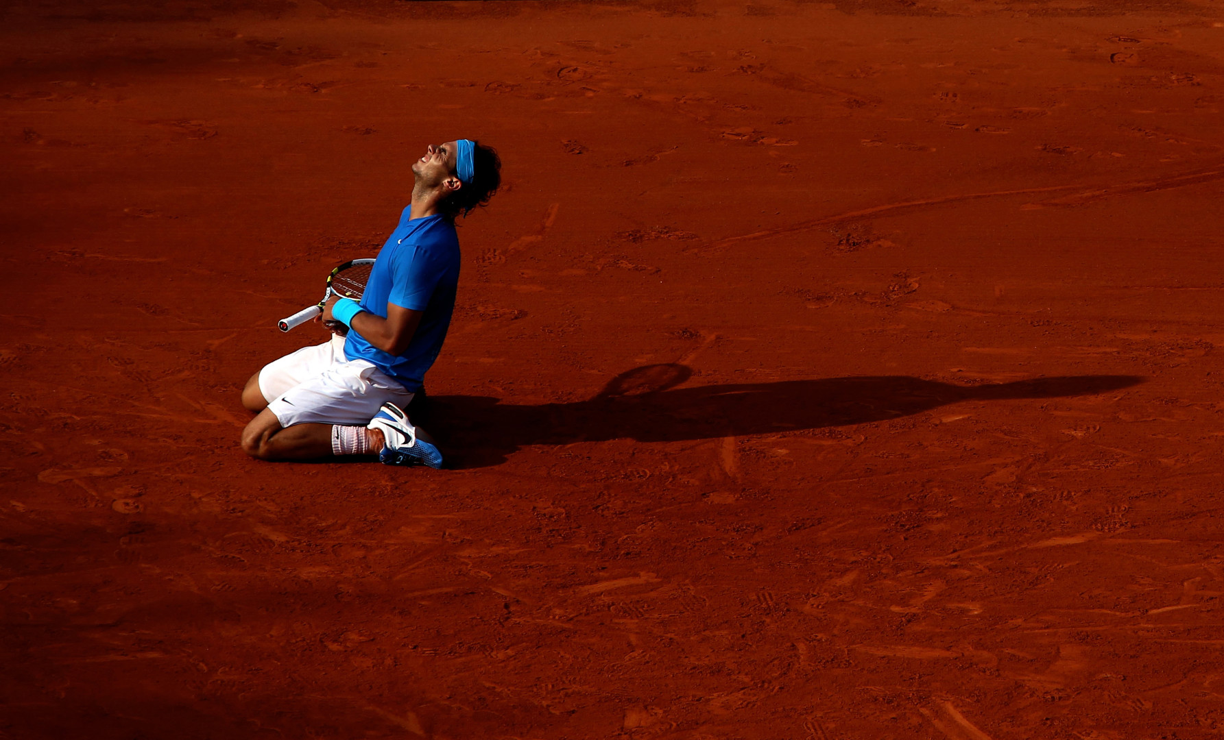 Rafael Nadal on his knees on clay court whilst clutching his tennis racquet at match point.