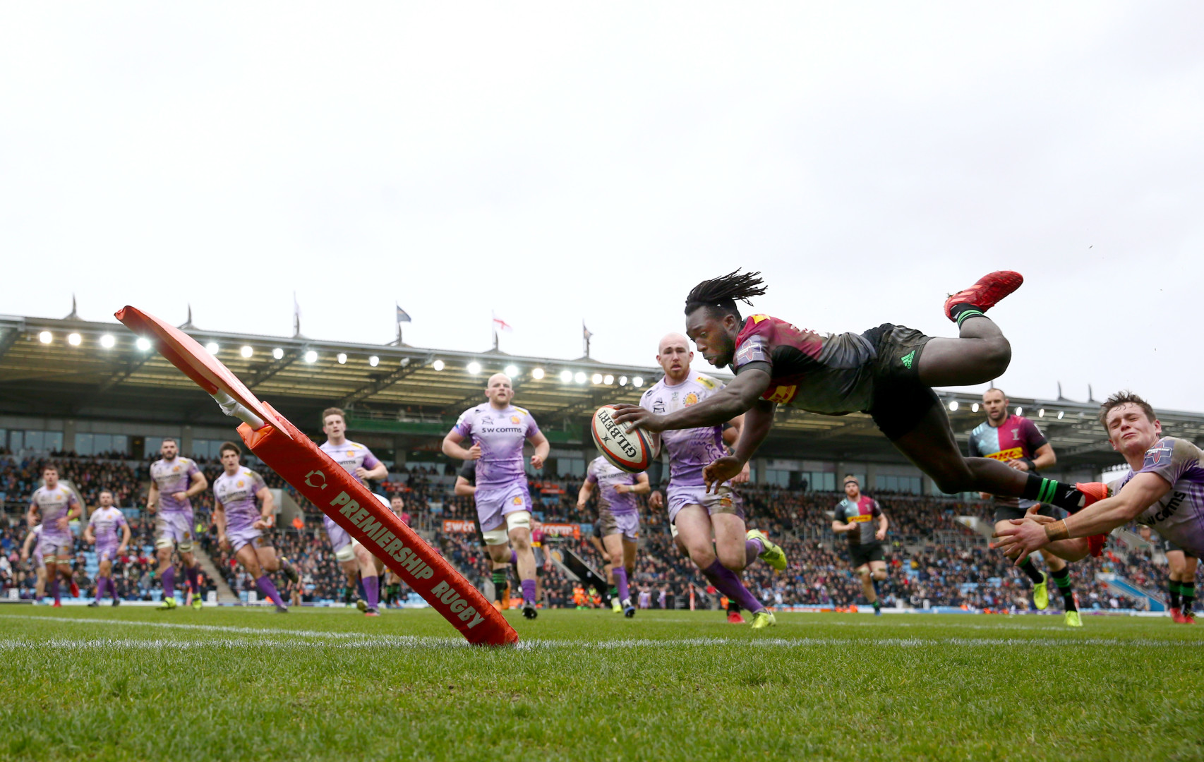 Harlequins player diving for a try in Rugby match as line of Exeter Chiefs players watch.