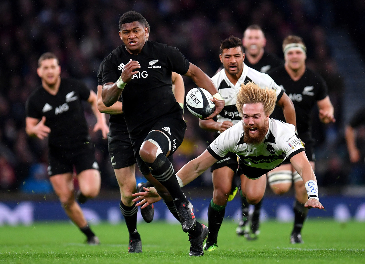 All Blacks rugby player striding down the pitch as opposing player dives to catch his feet.