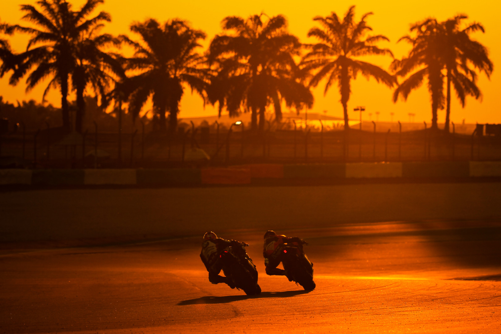 Two motorcyclists racing around a track bend in sync during sunset.