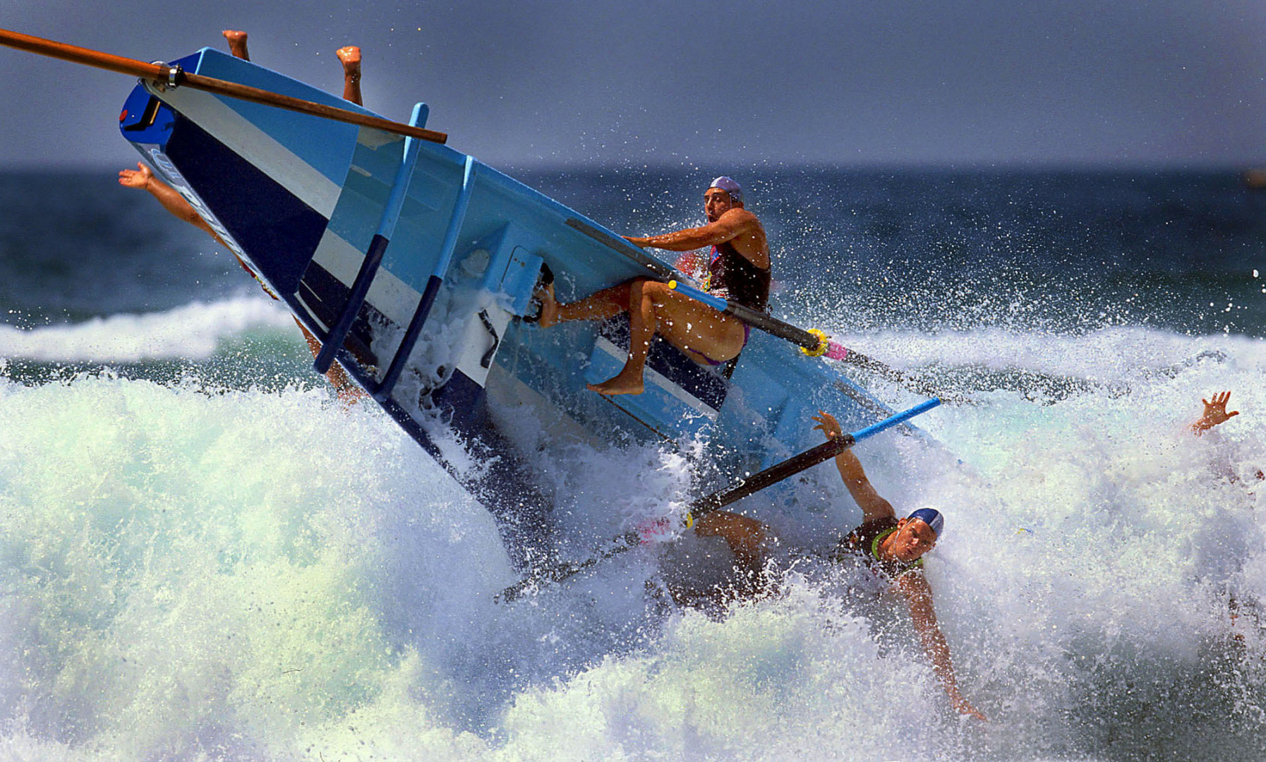 Offshore rowing boat capsizing after being wiped out by big wave whilst rowers are thrown from boat.