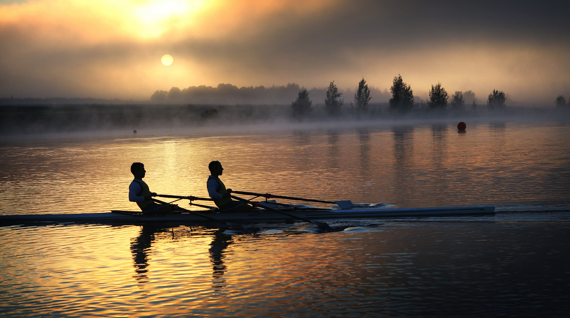 Two rowers practising rowing across a misty lake at sunrise.