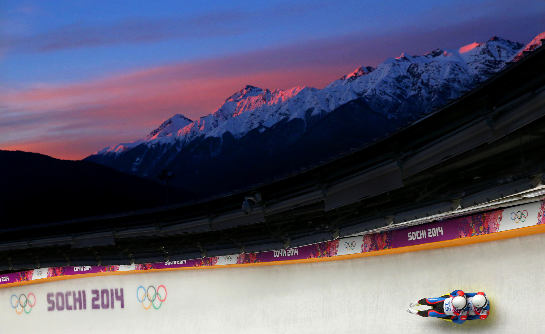 Two-man bobsleigh team sliding down track as sun sets over snowy mountains at the 2014 Sochi Winter Olympic games.
