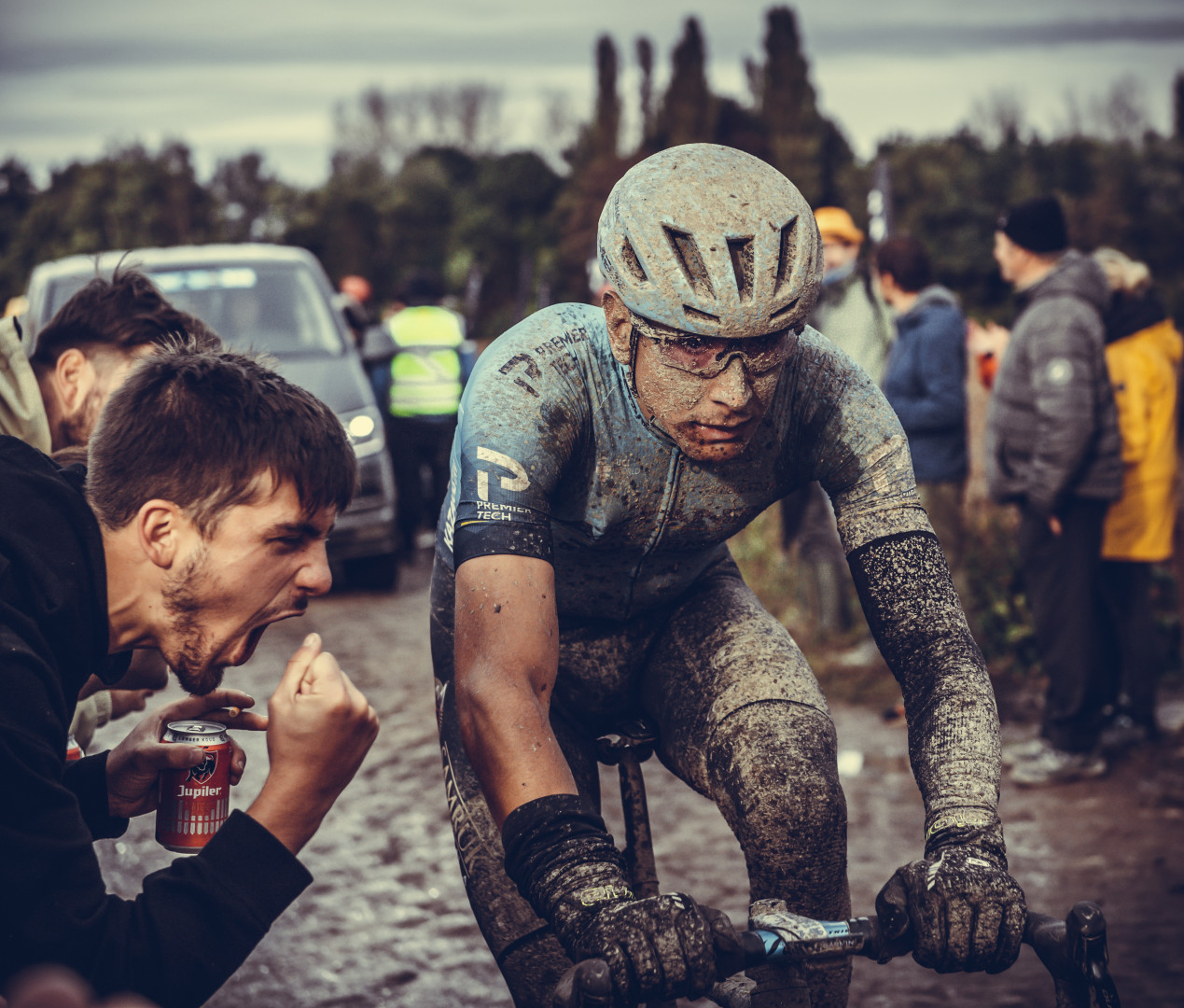 Muddy road cyclist at the 2021 Paris Roubaix as an enthusiastic man in the crowd shouts at him.
