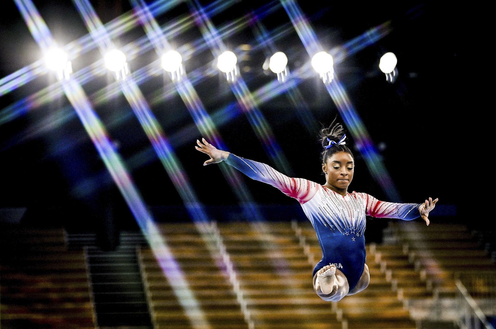 Simone Biles performing a split jump in beam routine as lights shine down behind her.