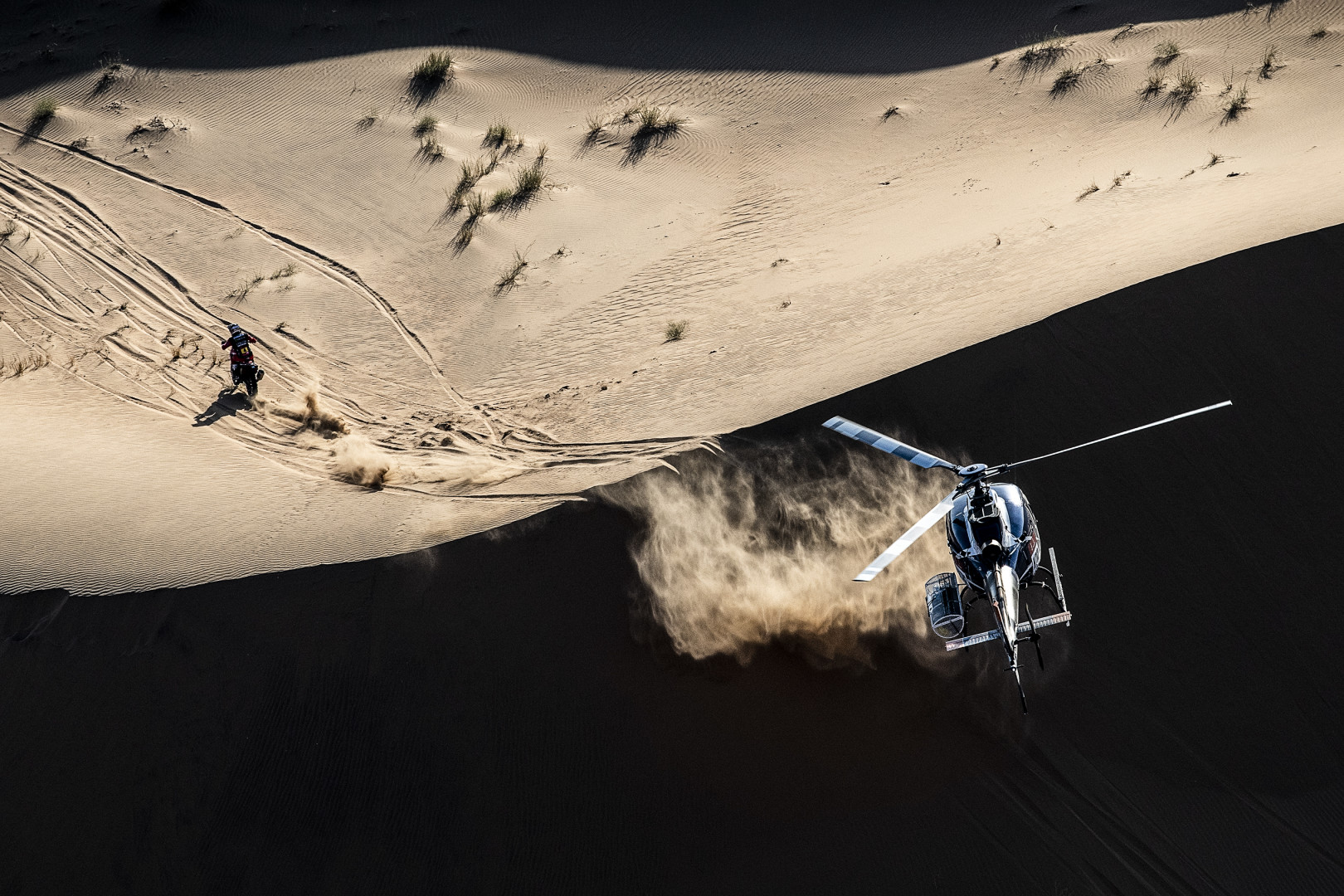Helicopter follows motorcyclist over sand dunes in Dakar Rally.