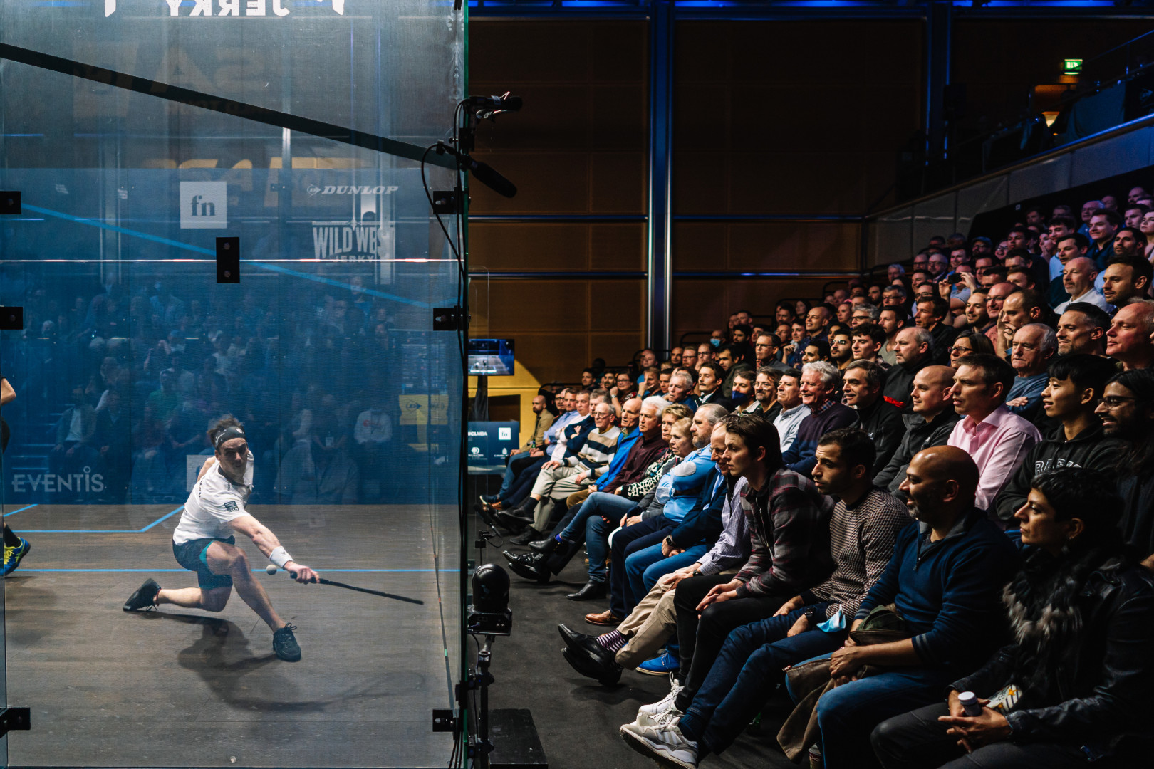 Split screen image of crowd watching squash player about to hit a shot at the Canary Wharf Squash Championship 2021.