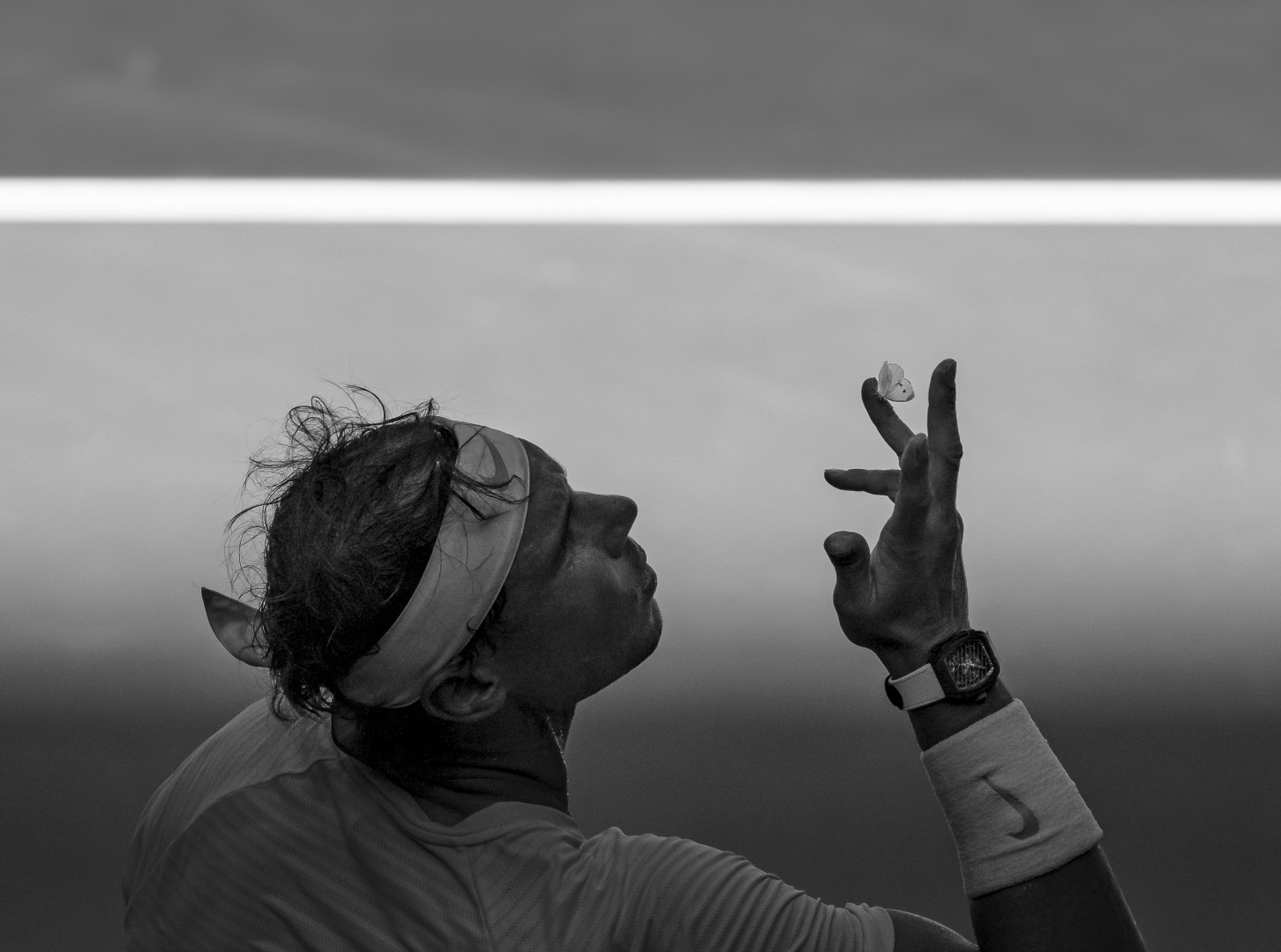 Black & White image of Rafael Nadal blowing a butterfly off his fingers whilst sitting on the side lines.