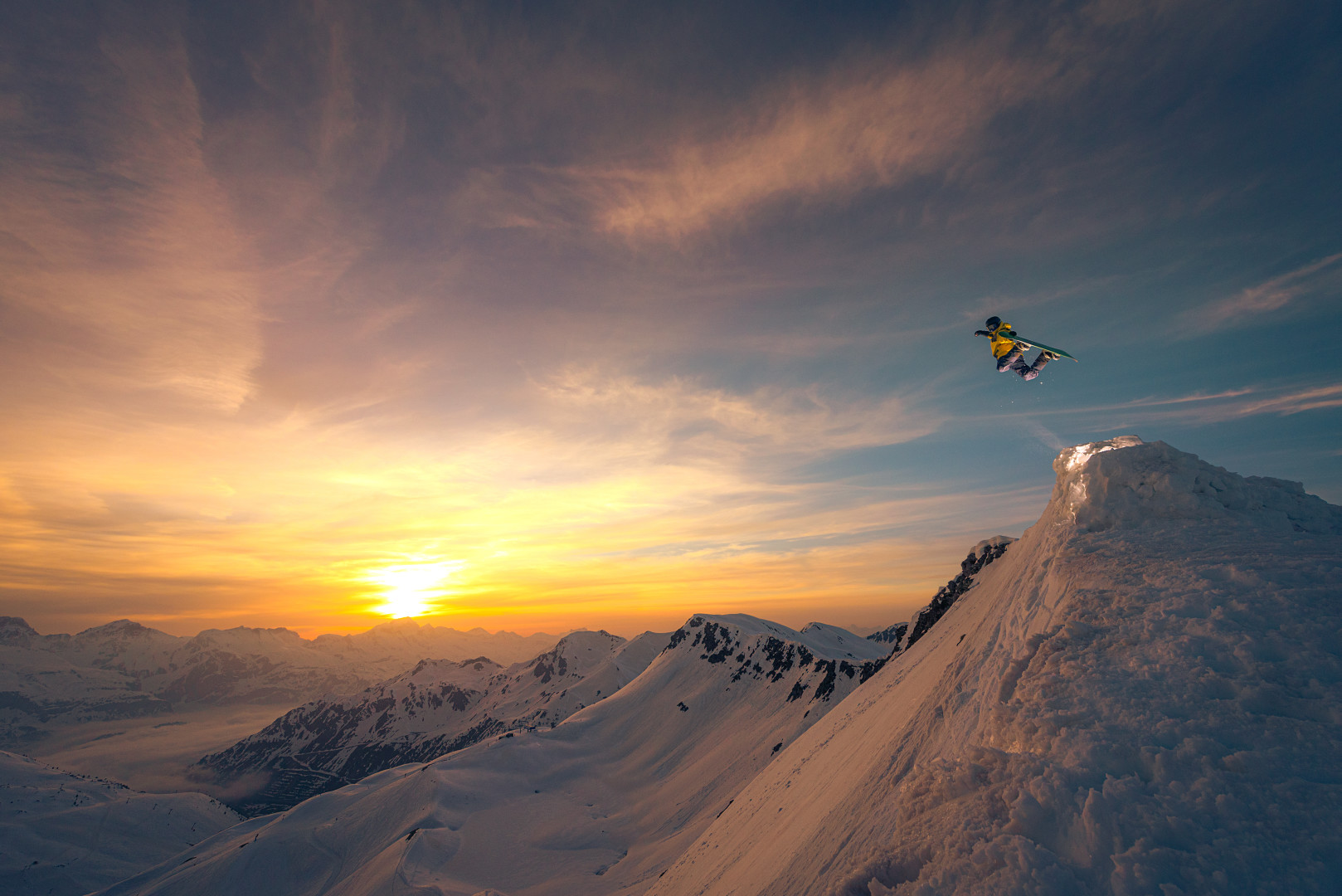 Snowboarder flying of peak of mountain top as the sun sets in the distance.