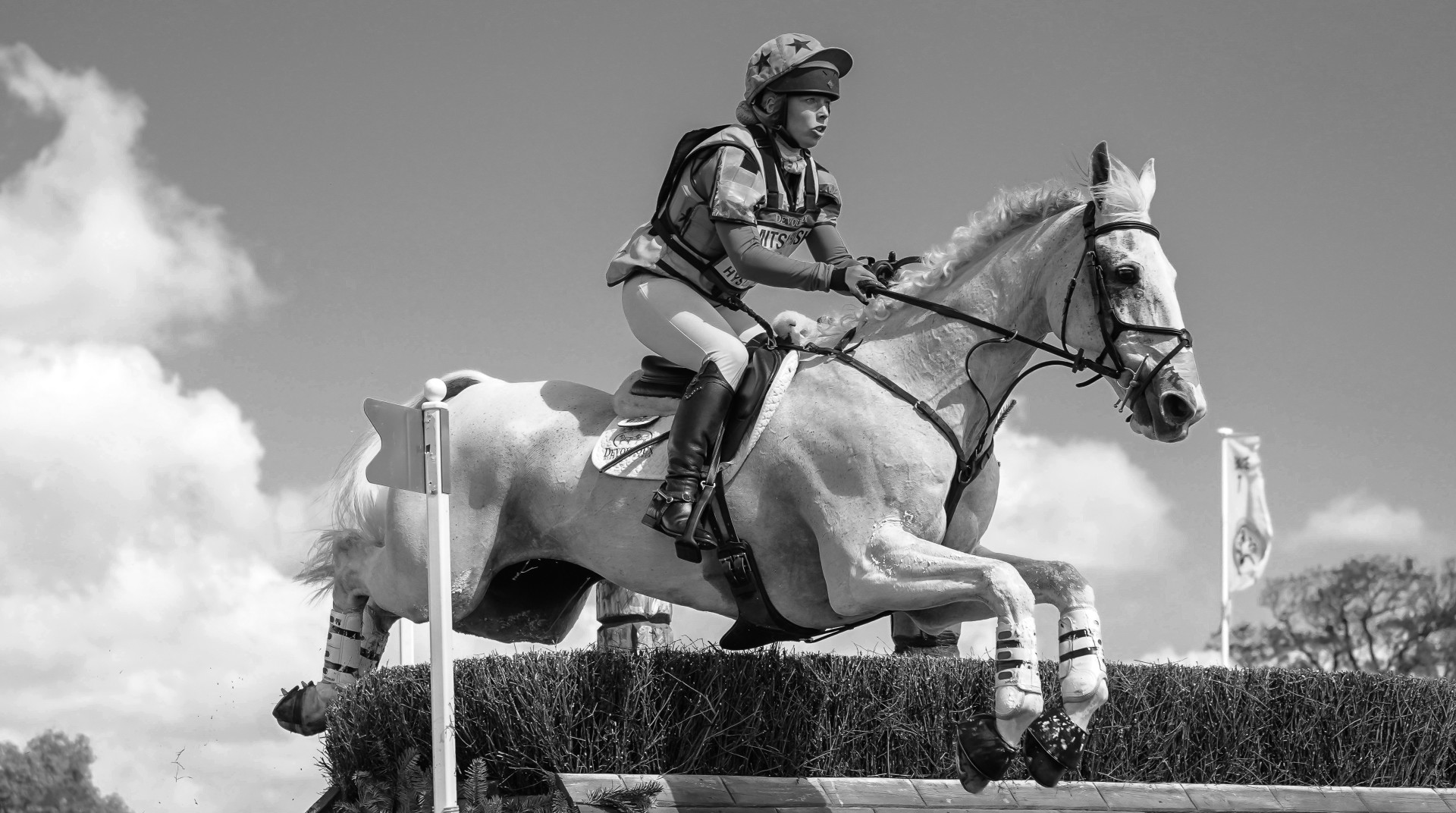 Black & White photograph of female jockey jumping a hedge hurdle on her white horse.