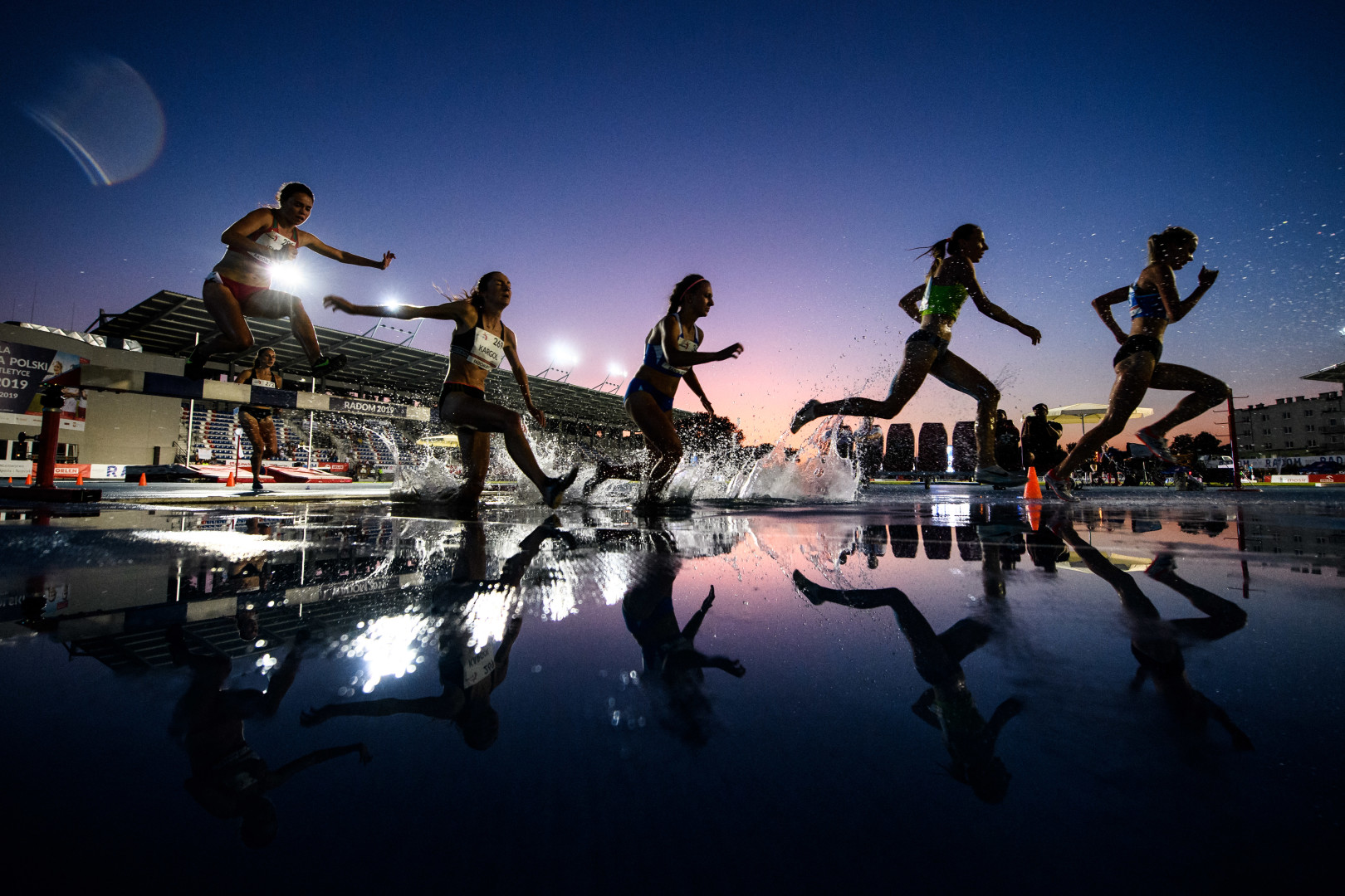 Female athletes landing in the steeplechase water as they clear the hurdle during an evening race.