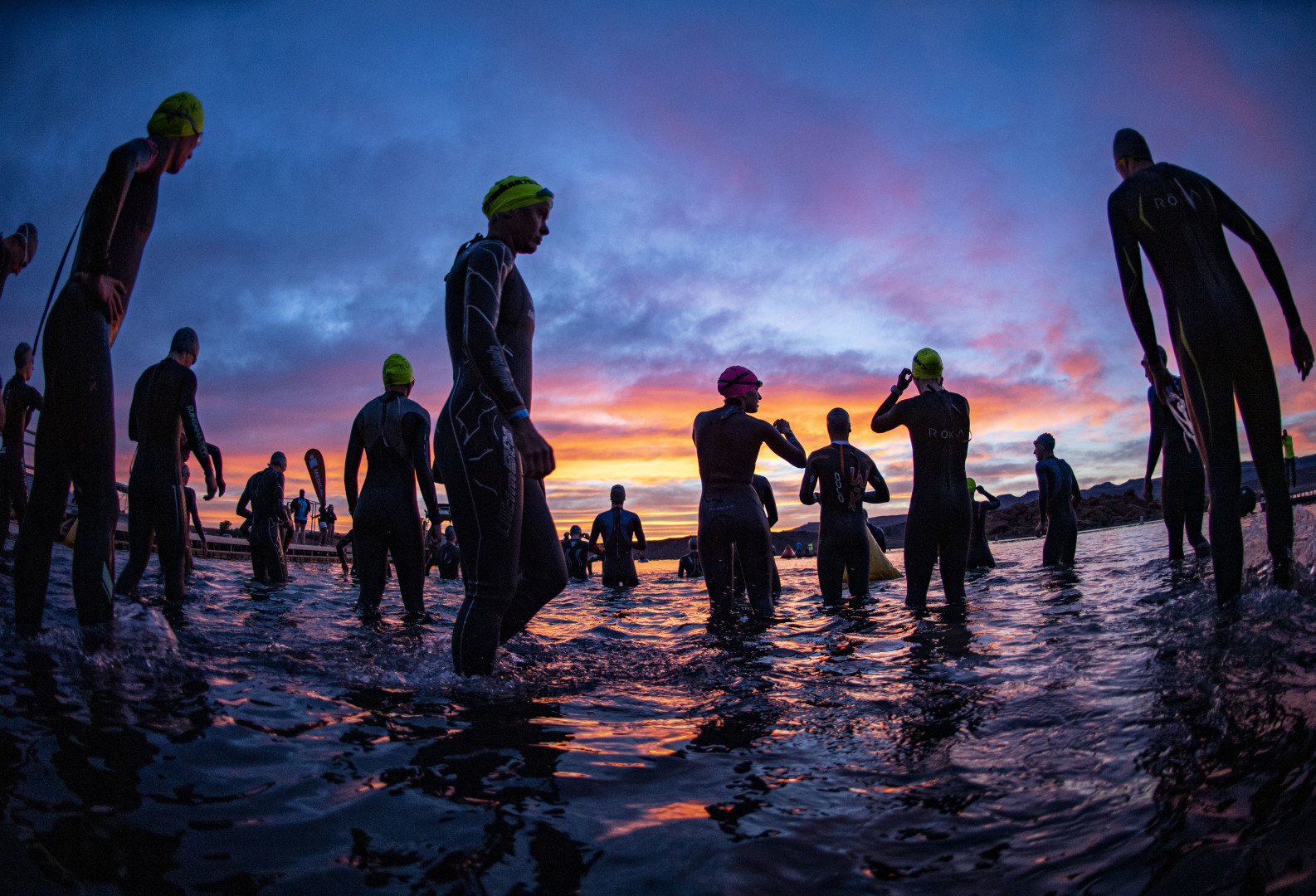 Cold water swimmers in wetsuits and swim caps entering water at a lake at sunrise.