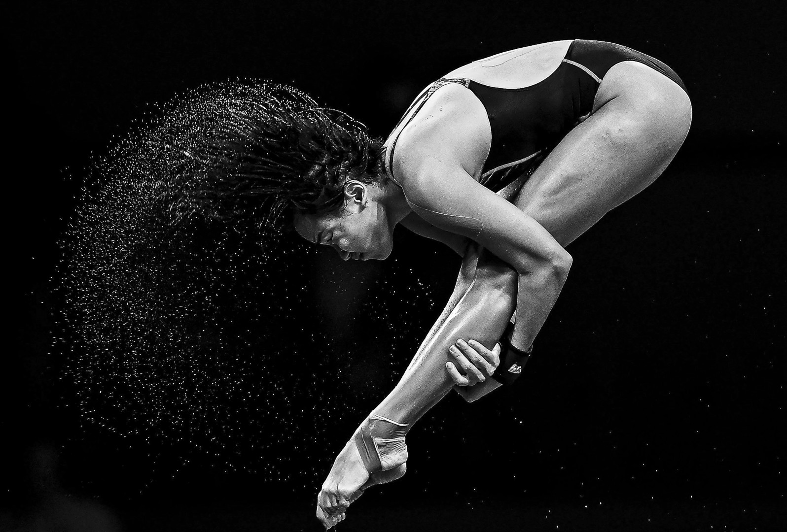 Black & White photograph of female diver in pike position, mid dive, as water sprays from her hair.