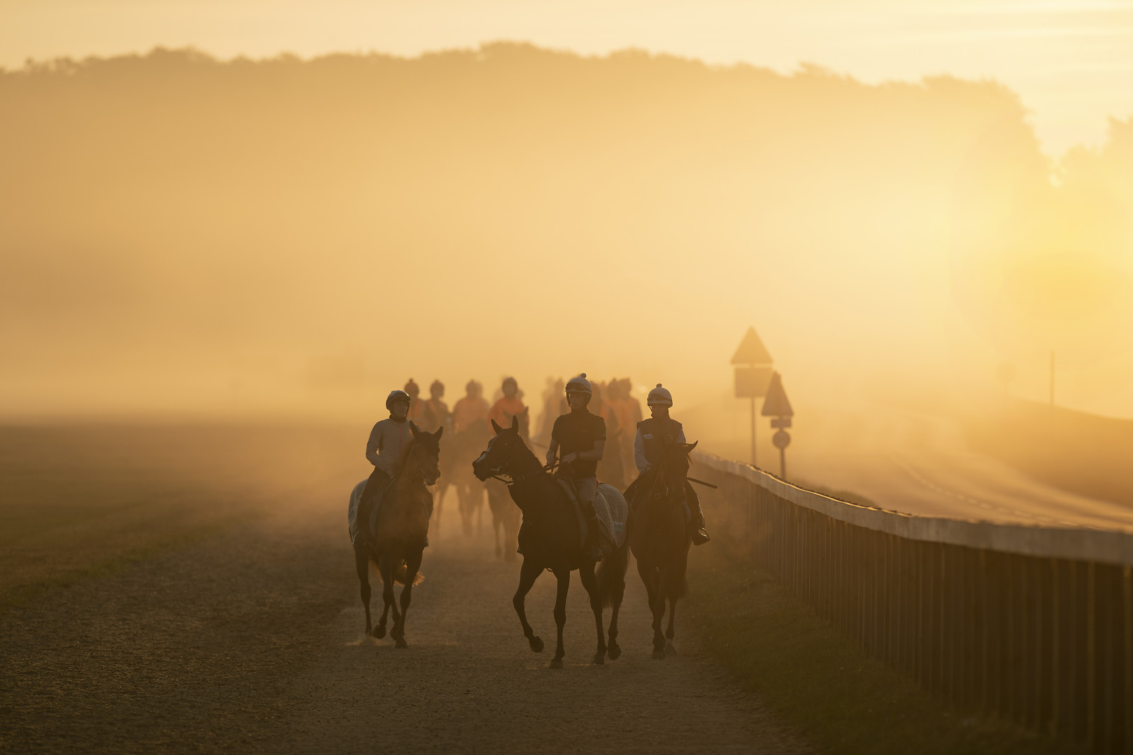 Group of jockeys slowly riding their horses during a misty morning sunrise.