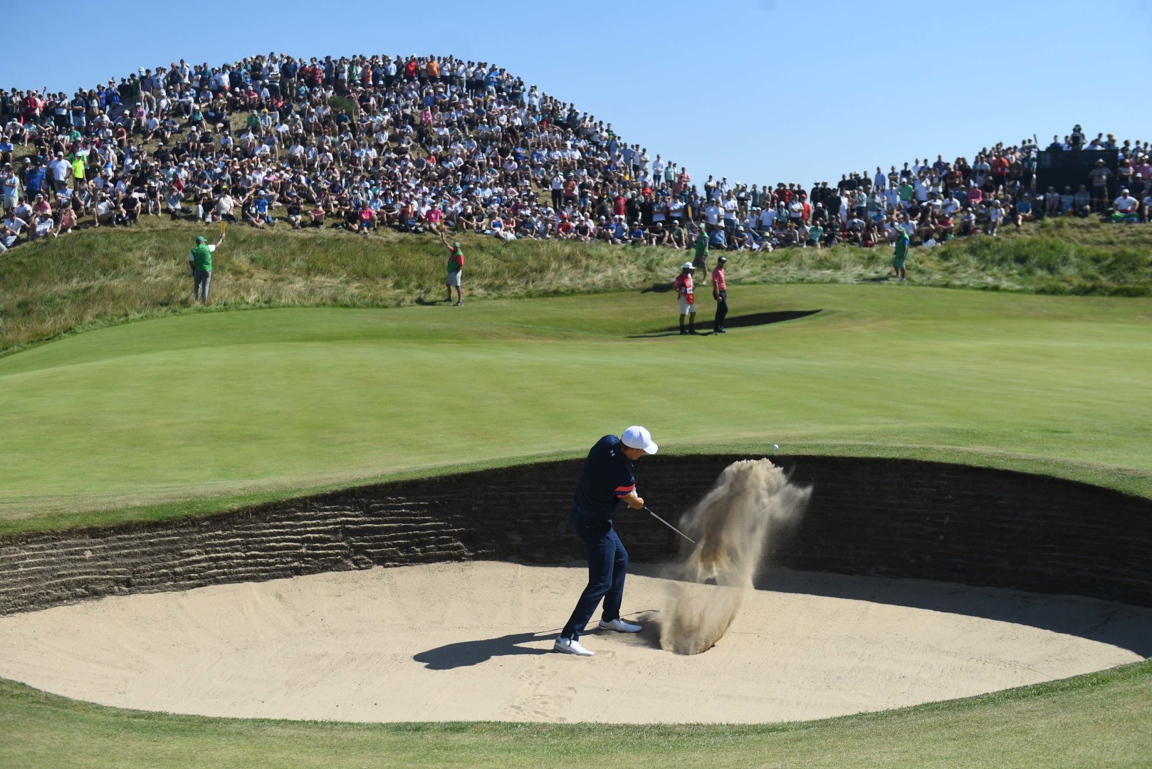 Male golfer hitting ball out of the deep sand bunker as wavy hill full of crowds of people watch on.