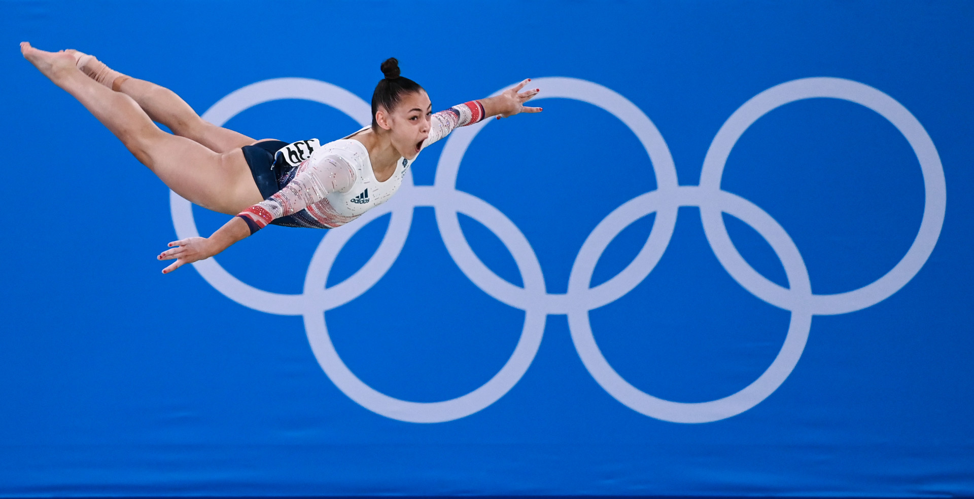 Gymnast flying across the gym floor like a bird as the Olympic rings sit in the background.