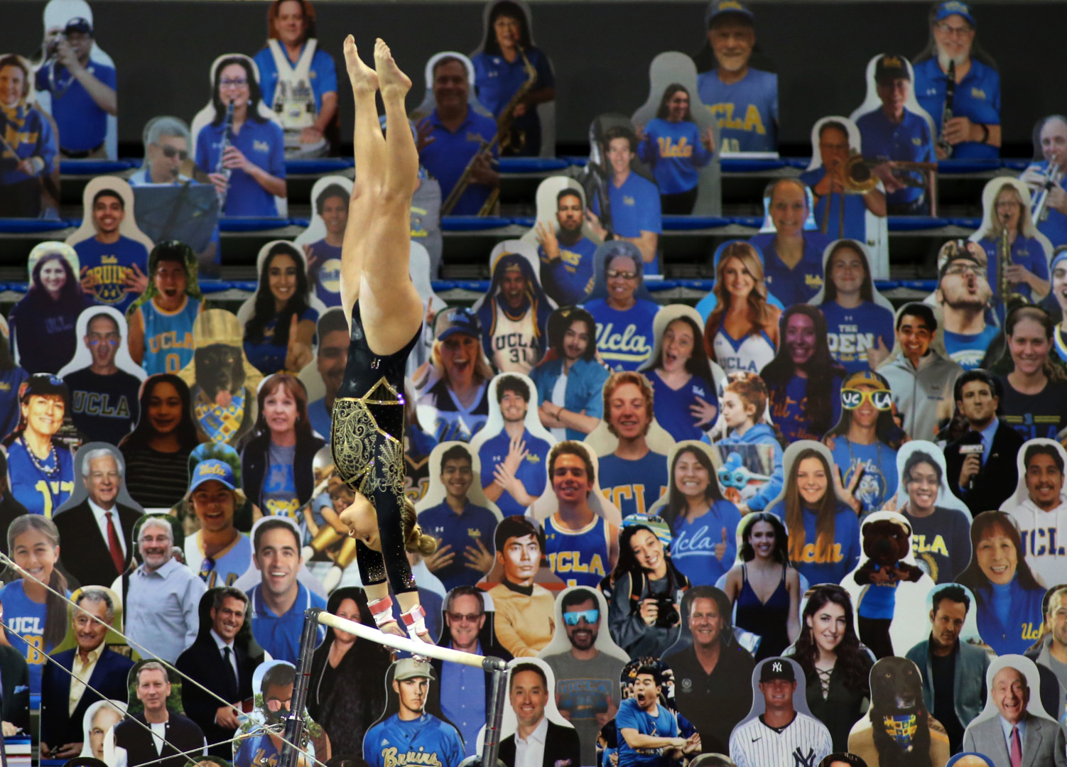 Female gymnastics performing on the swing bars as a crowd of cardboard cut-out people fill the stands at the Tokyo 2020 Olympics.