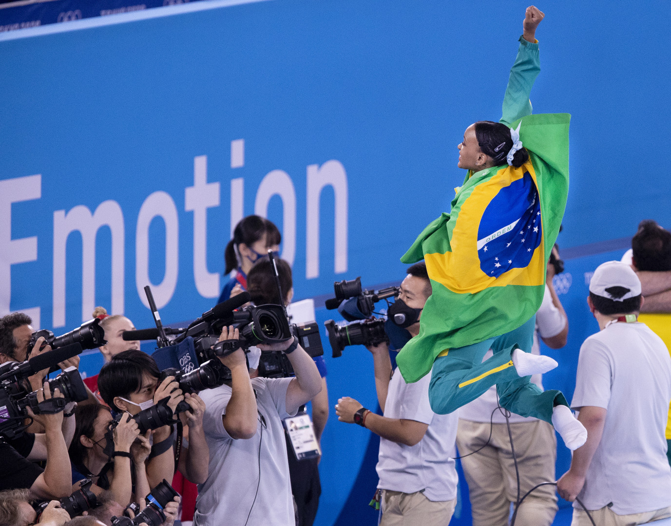 Rebecca Andrade celebrates winning gold by jumping in the air with the Brazilian flag around her shoulders as photographers take photographs.