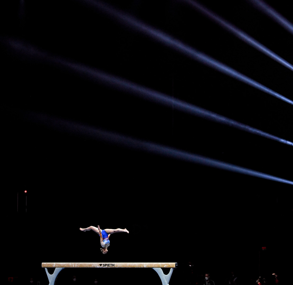 Gymnast upside down in the splits over the beam as light rays beam across the arena.