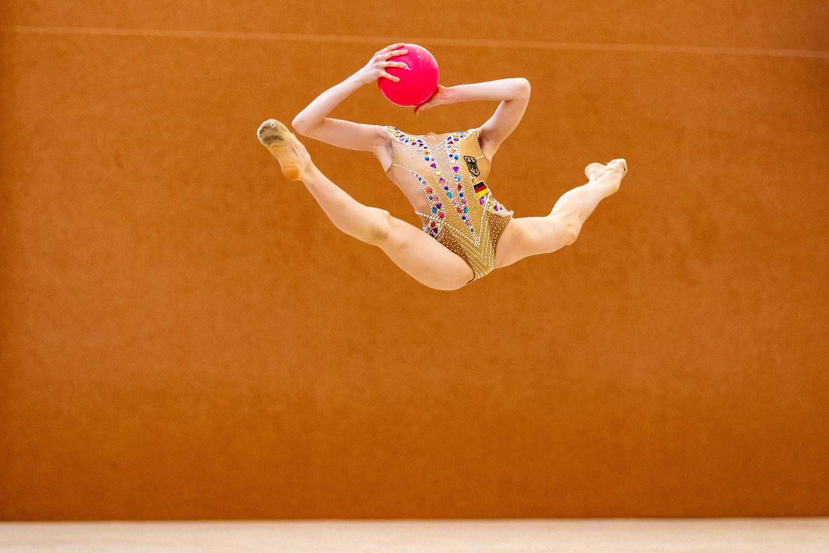 Rhythmic gymnastics mid air in the splits holding a ball in the position that her head should be.