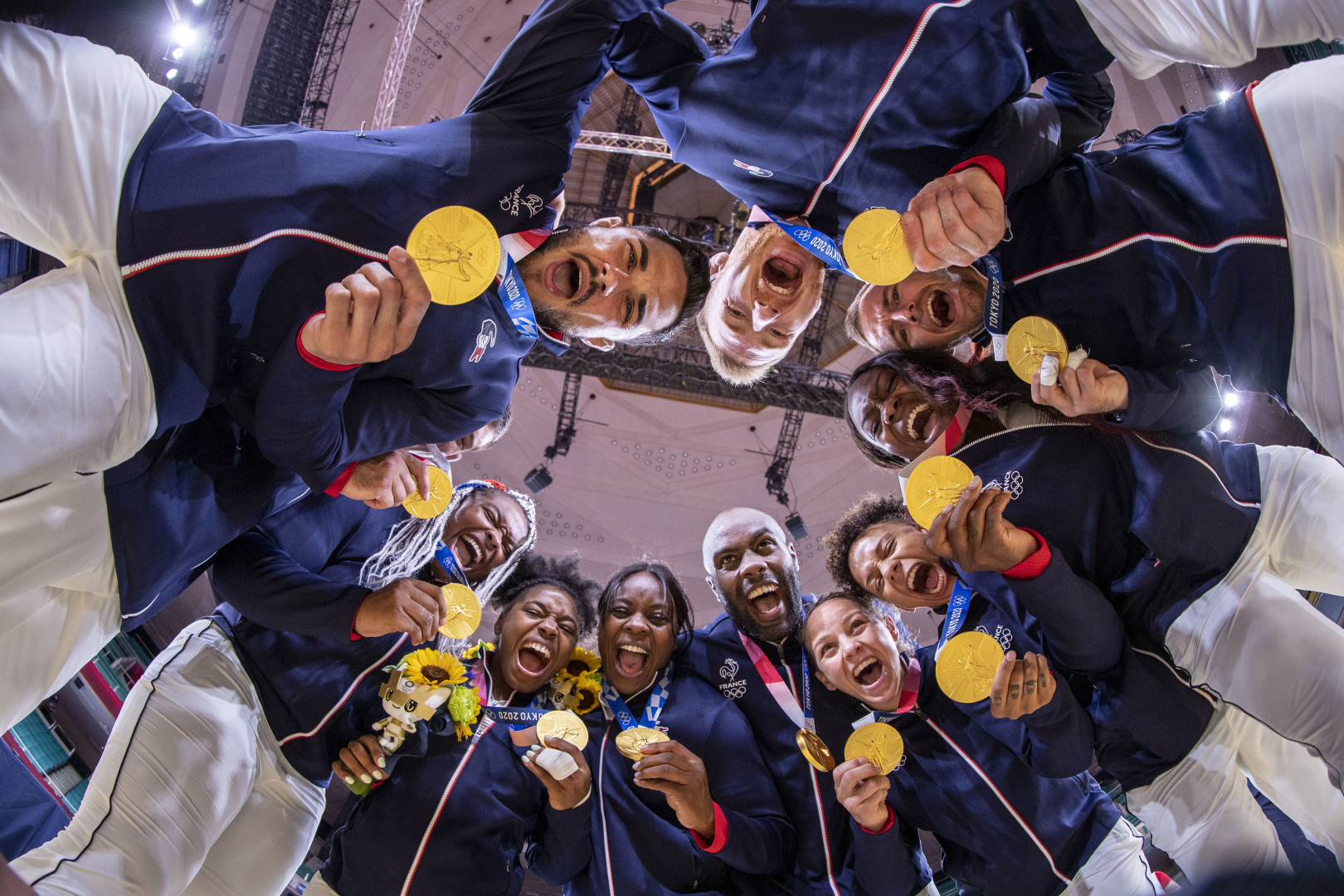 French Judo team huddled in a circle showing off their gold medals to the camera below them.