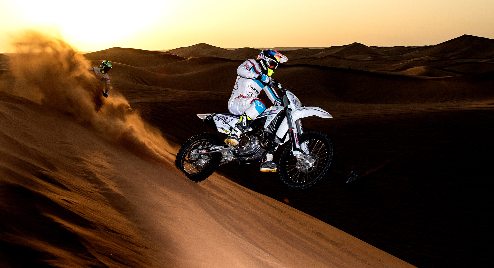 Motor cyclist riding on their back wheel down a sand dune whilst spraying the rider behind with sand.