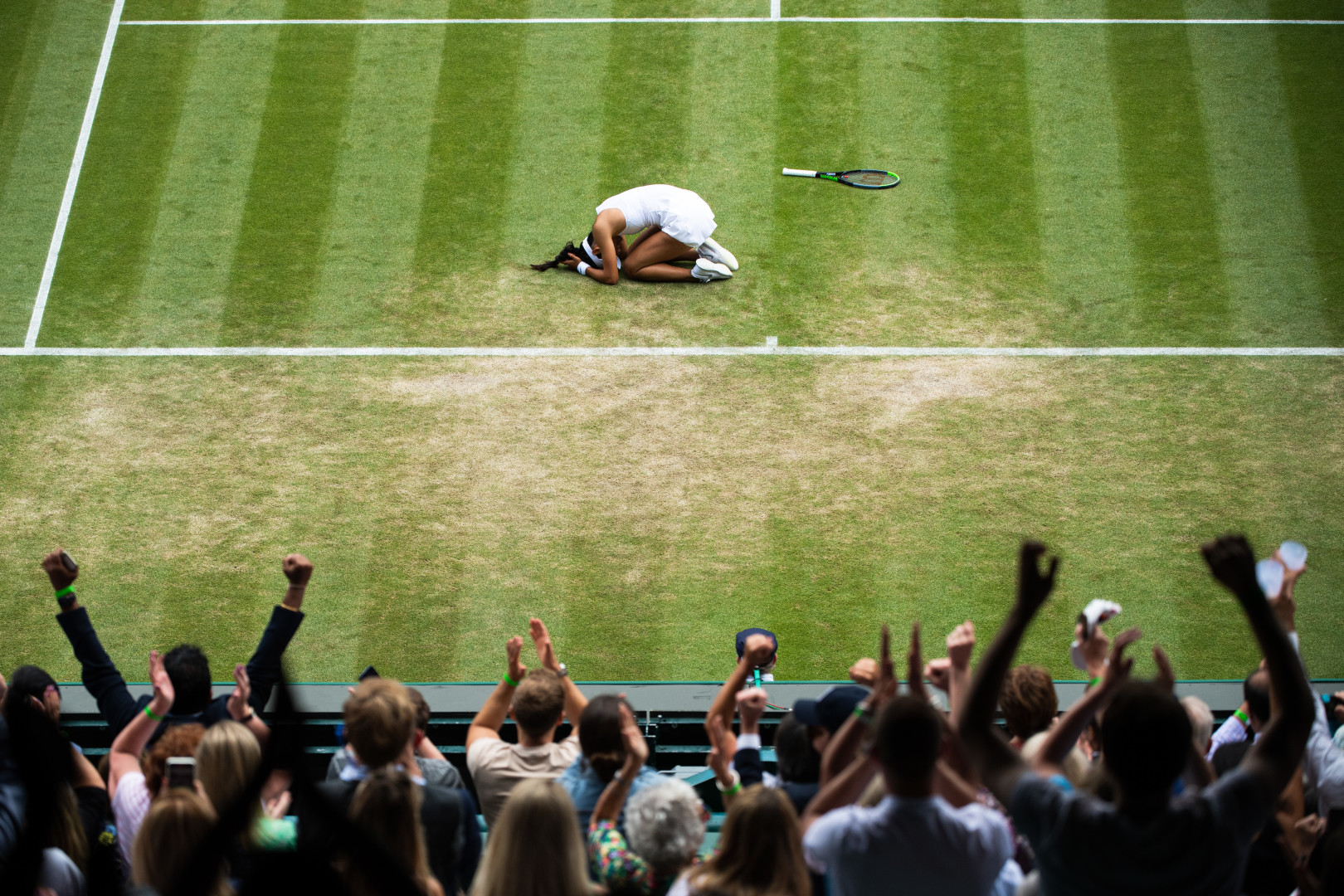 Female tennis player in a bawl on the court floor as crowd celebrates her win at the Wimbledon Tennis Championships.