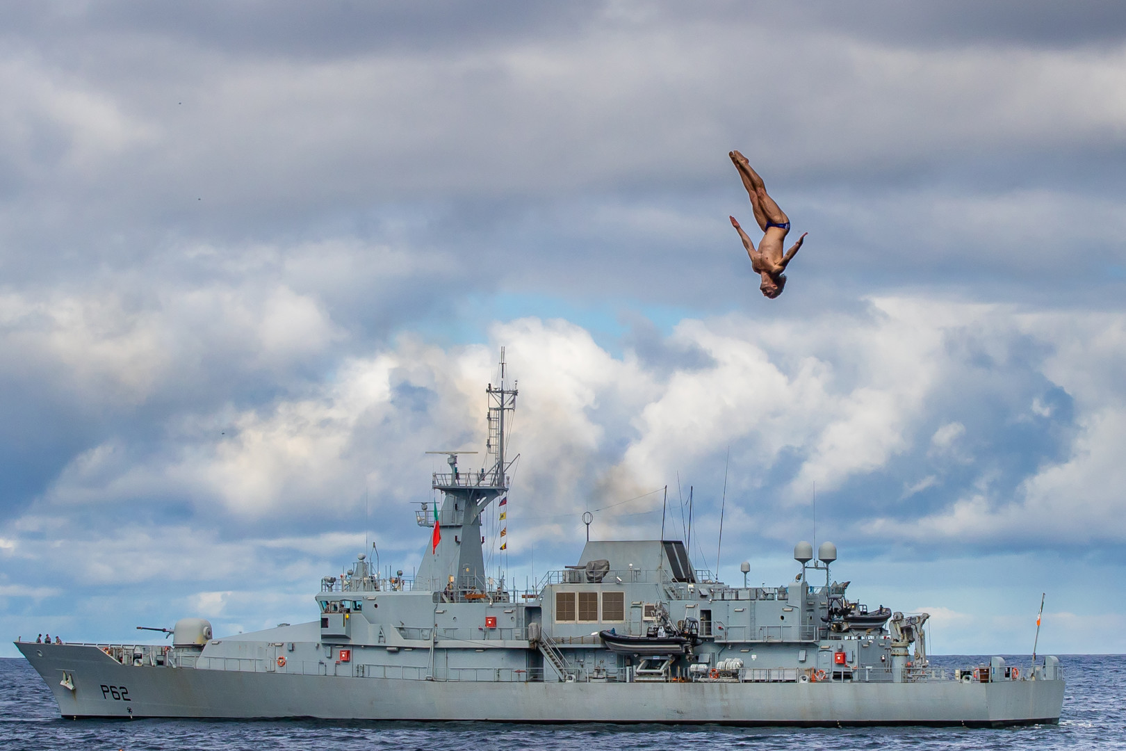 Cliff diver mid air as Naval ship sits in the distance at sea.