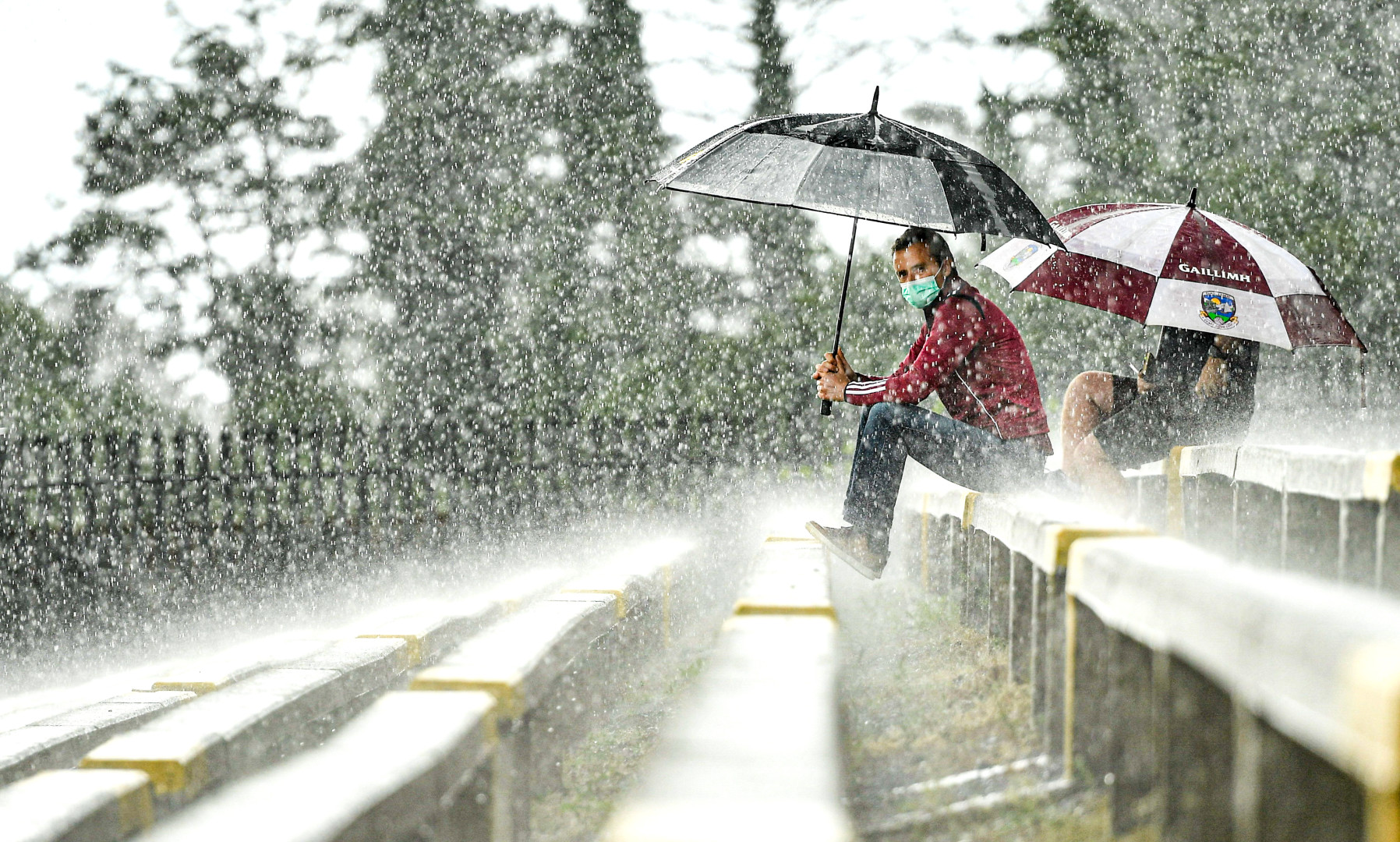 Coaches sitting on the open stands under umbrellas in the rain at a Gaelic football ground.